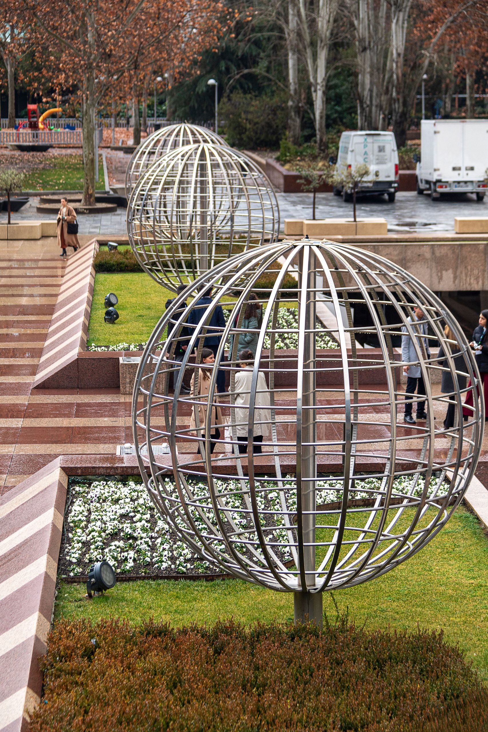 Metal sphere sculptures in a Madrid park with people walking nearby on a cloudy day.