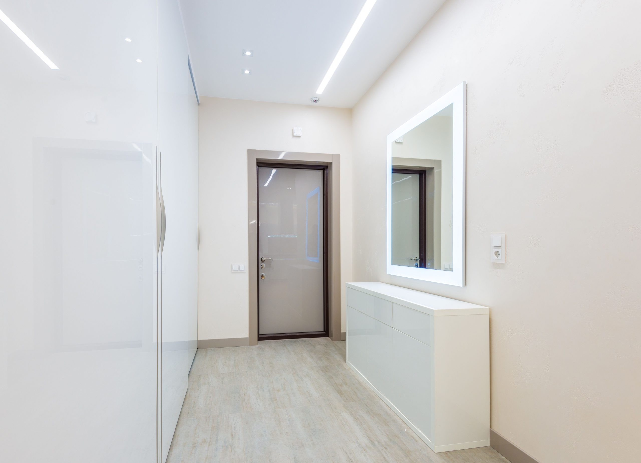 Minimalistic interior design of light hall of tall spacious closet near glass door with wooden frame and opposite white cabinet under led mirror in corridor