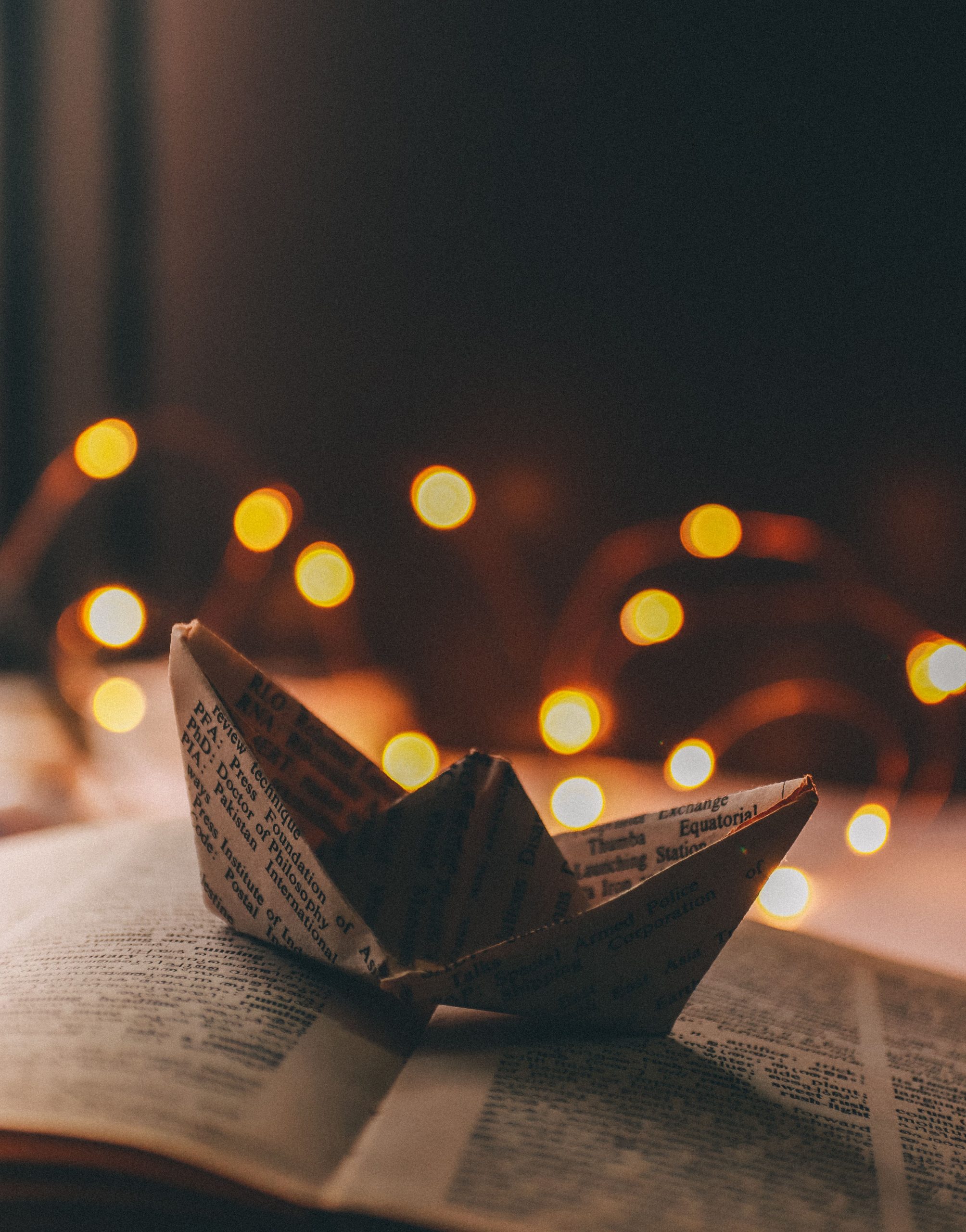 Origami ship made with book sheet placed on open book with illuminated garland on background in light room