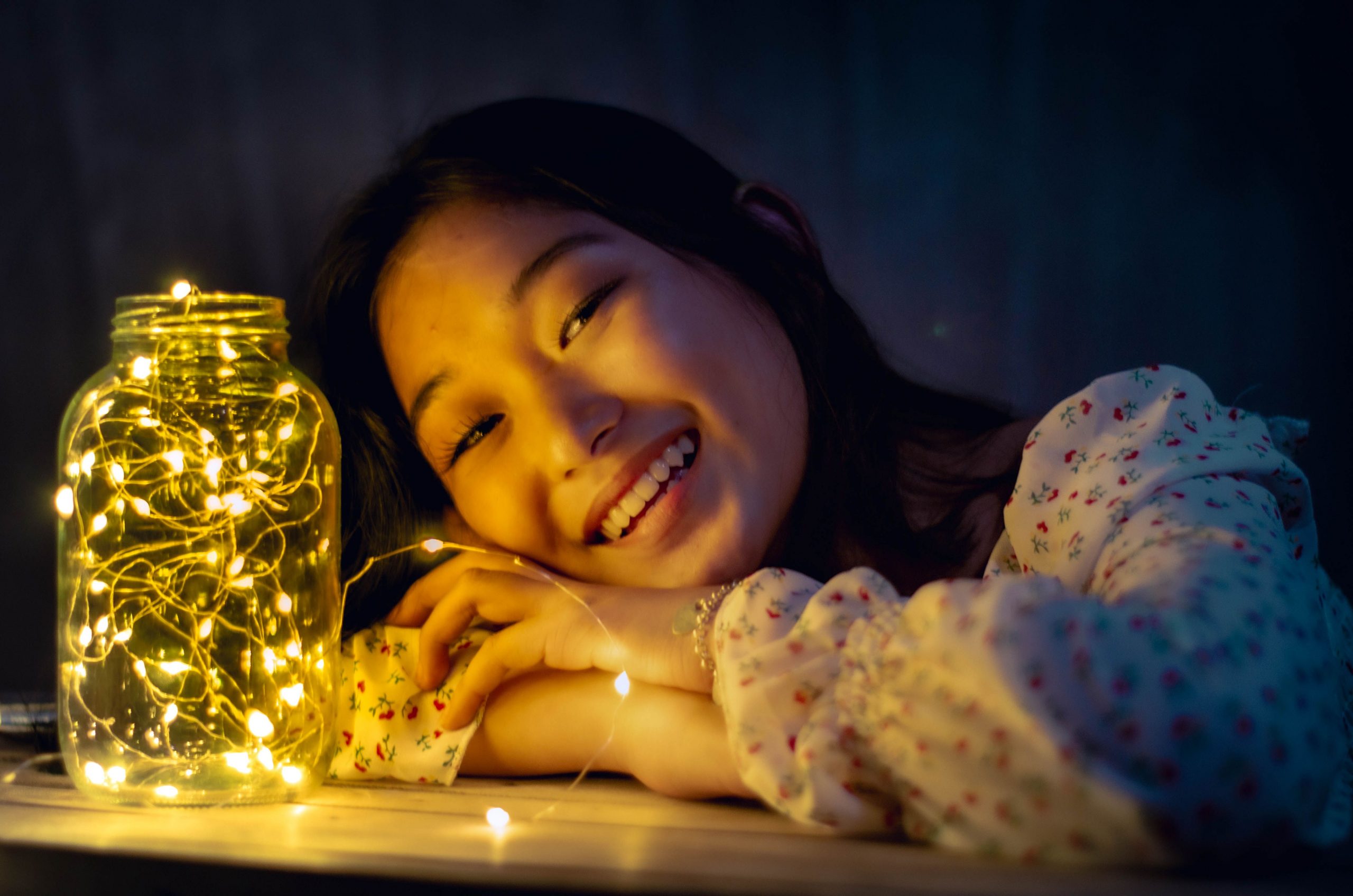 Smiling young woman resting her head near a jar with glowing fairy lights, creating a warm ambiance.