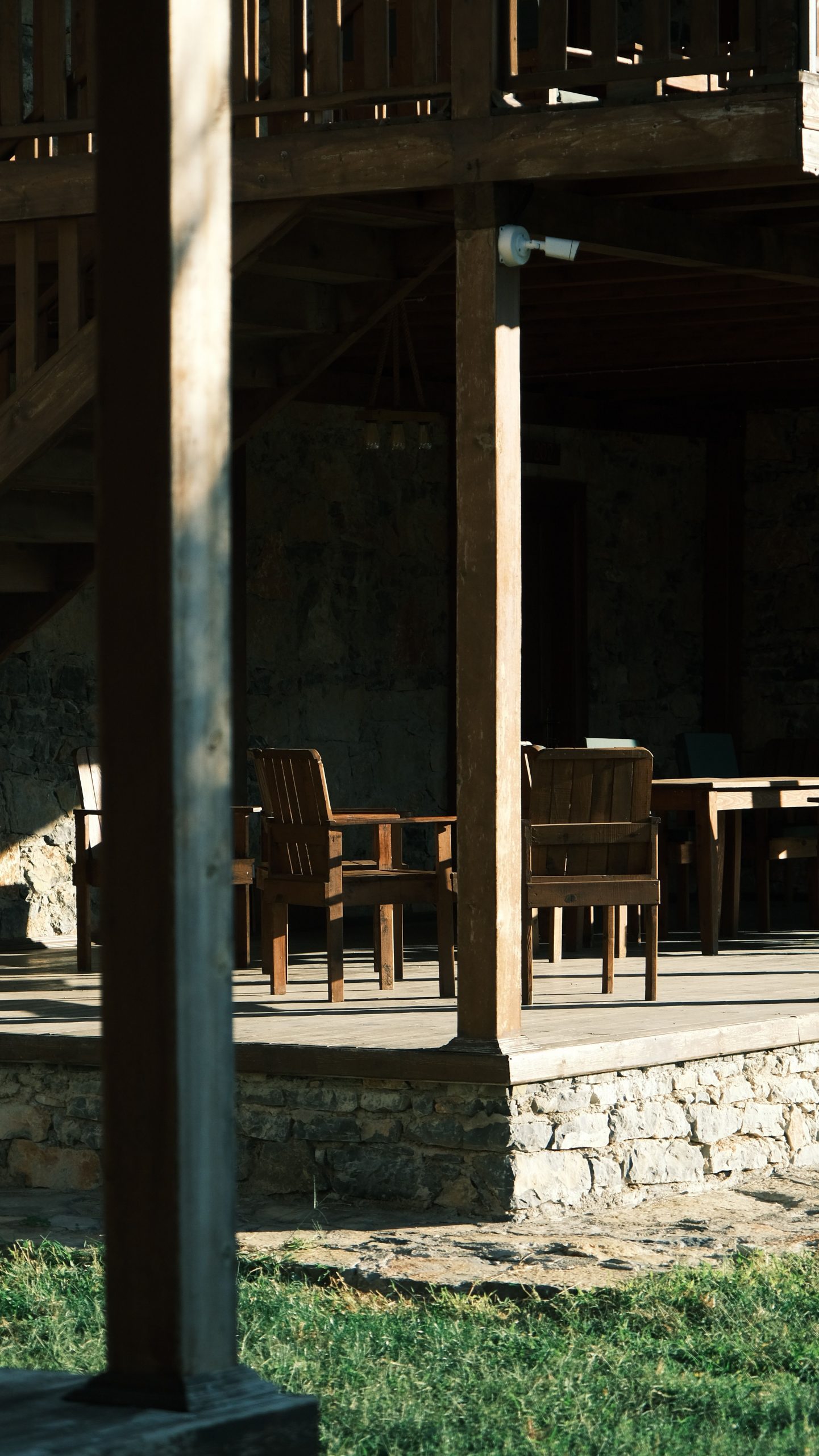 Sunlit wooden patio with rustic chairs by stone wall in serene setting.