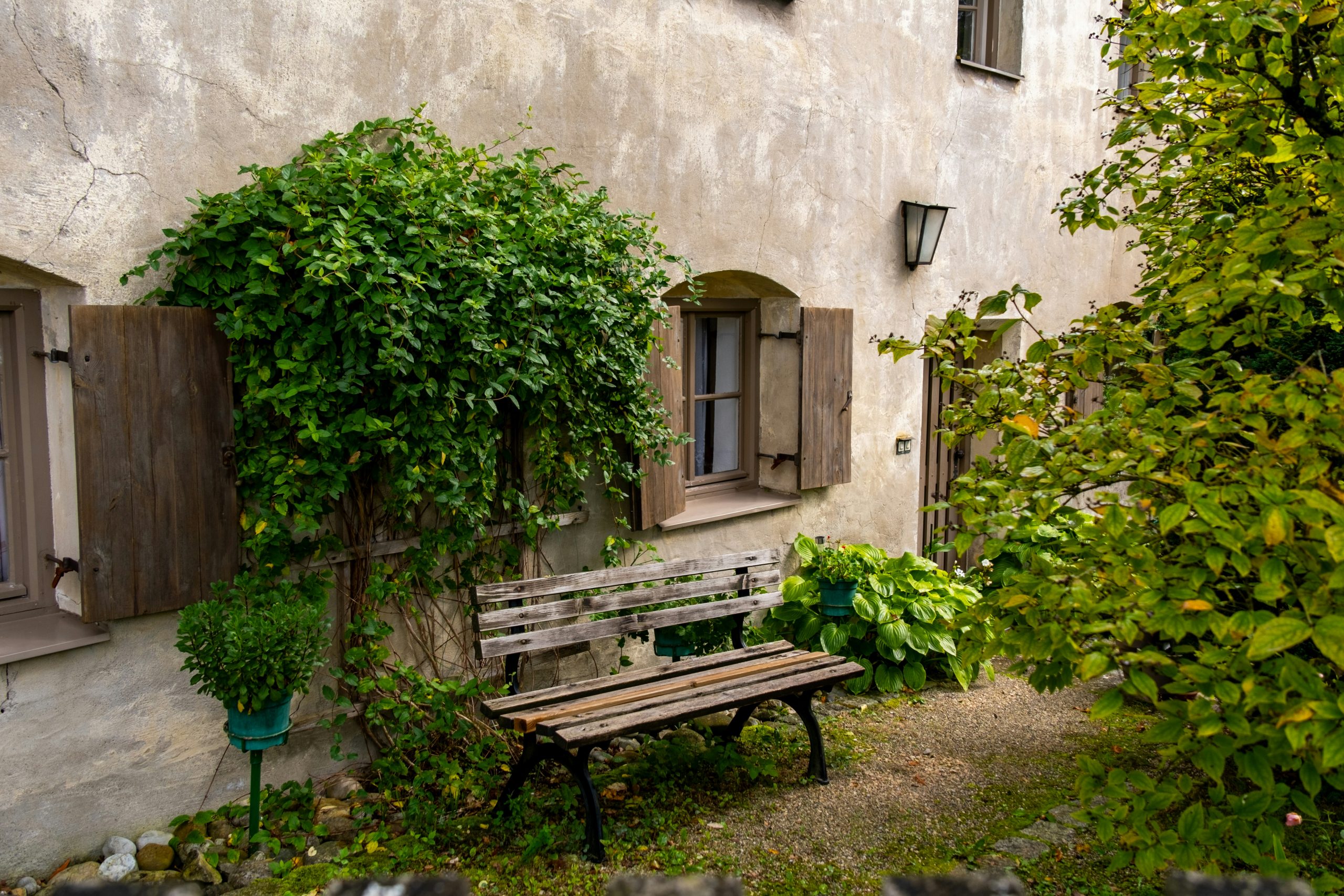 This image showcases a peaceful and picturesque courtyard with an old wooden bench placed in front of a rustic house. The weathered stone wall and wooden shutters give the house a vintage charm. A lus