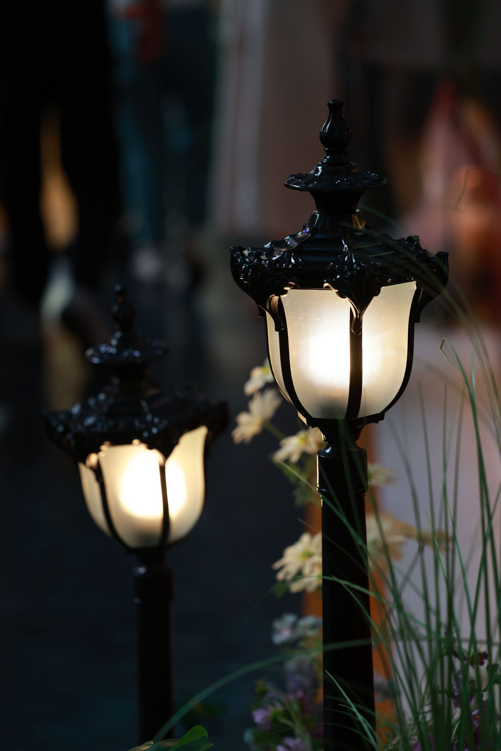 Two vintage-style street lamps illuminate a garden path at dusk, casting soft light.