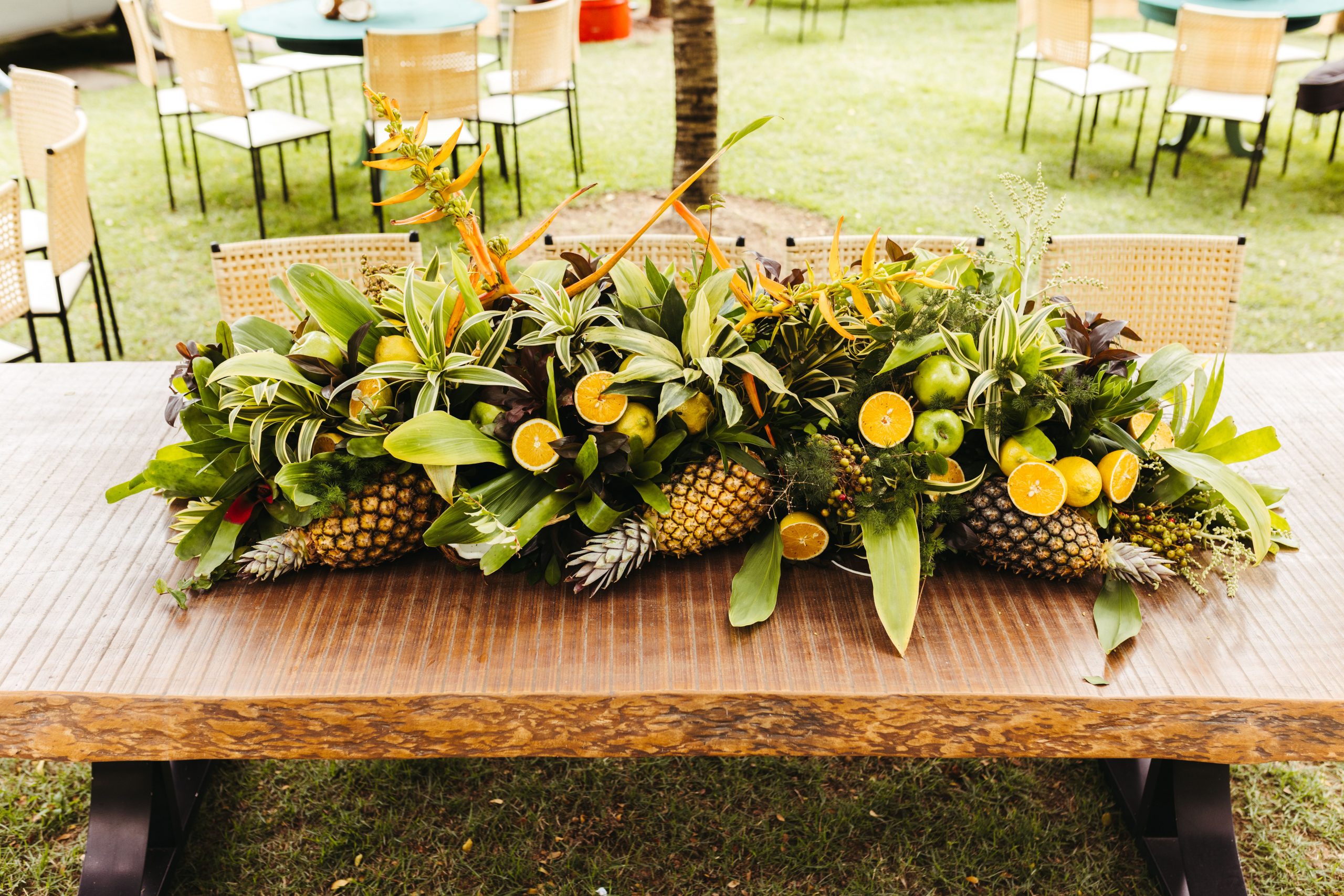 Vibrant tropical fruit and greenery centerpiece on a wooden table in an outdoor setting.