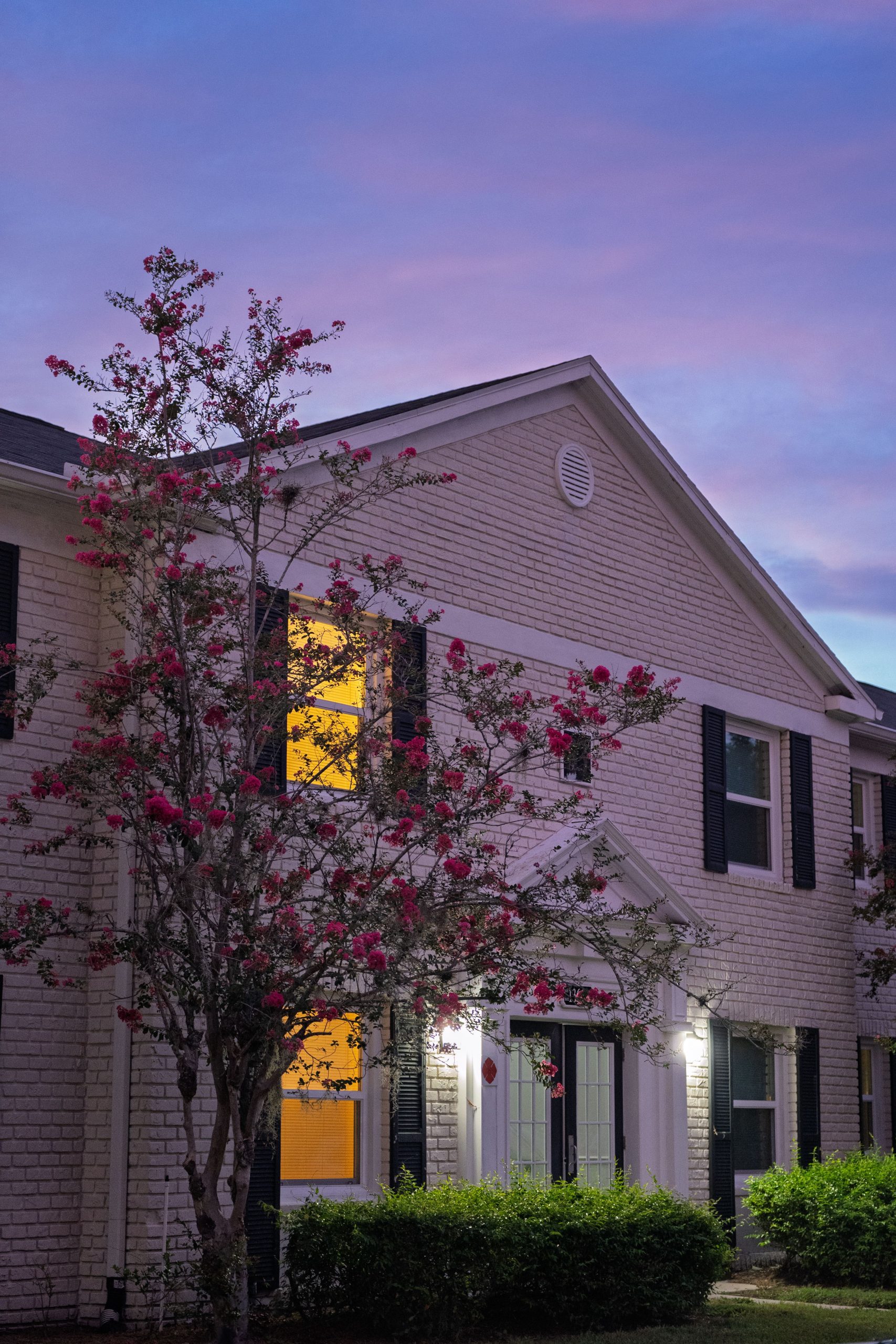 White brick suburban house with pink blossoms at twilight under a vibrant sunset sky.