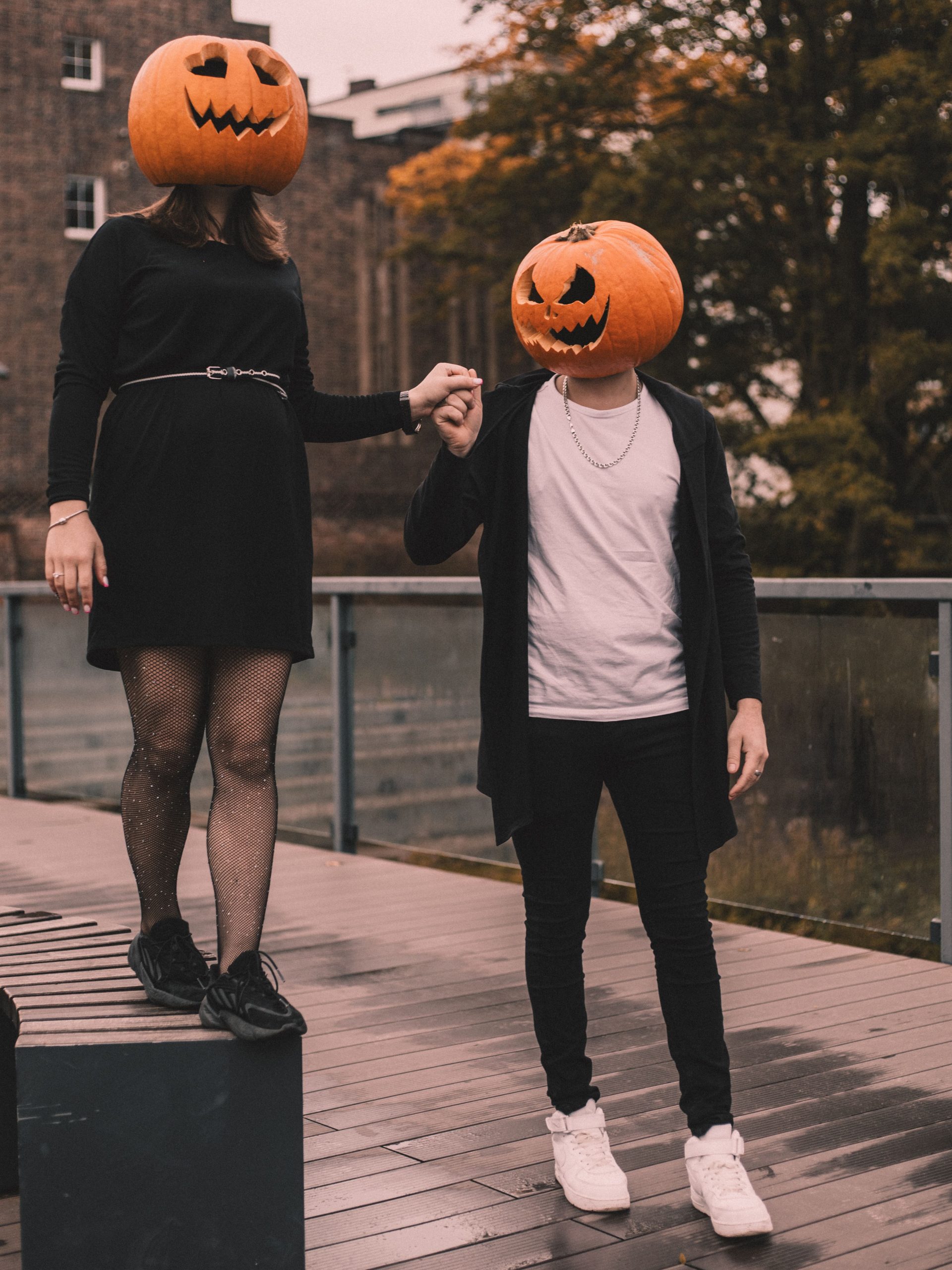 A couple dressed in black with pumpkin masks holding hands on a wooden deck, embracing Halloween spirit.