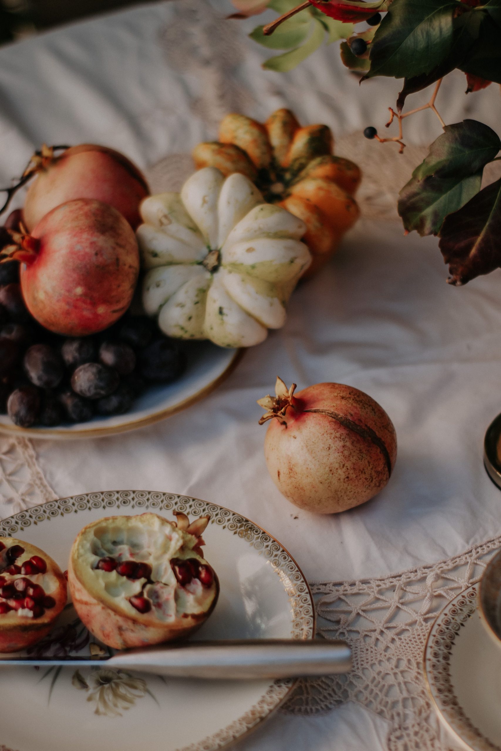A cozy autumn-themed still life featuring pomegranates, pumpkins, and grapes on a lace tablecloth.