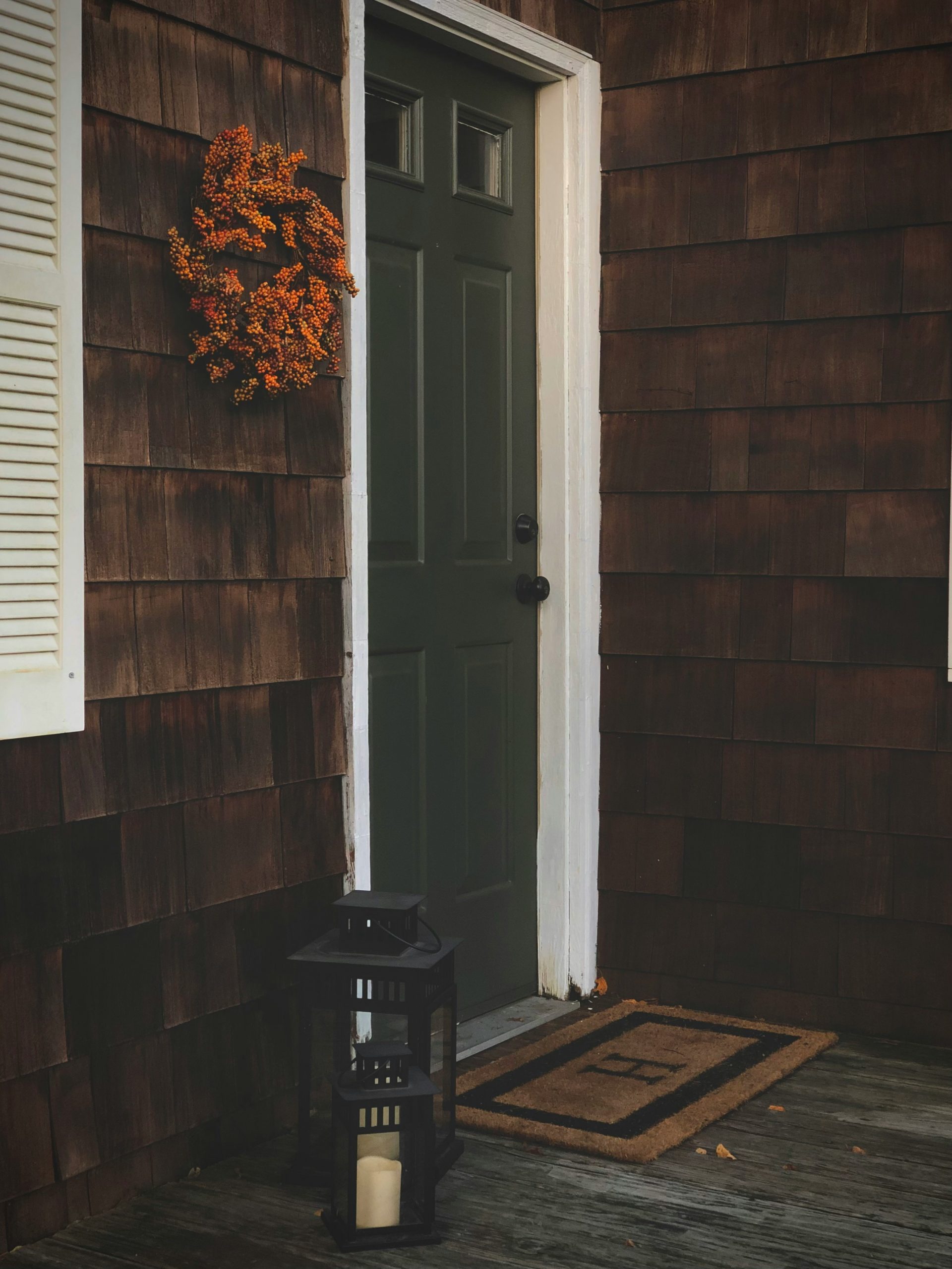 A green door with an orange fall wreath hanging to the left of it. Some black lanterns and a welcome mat with an H sits in front of the door.
