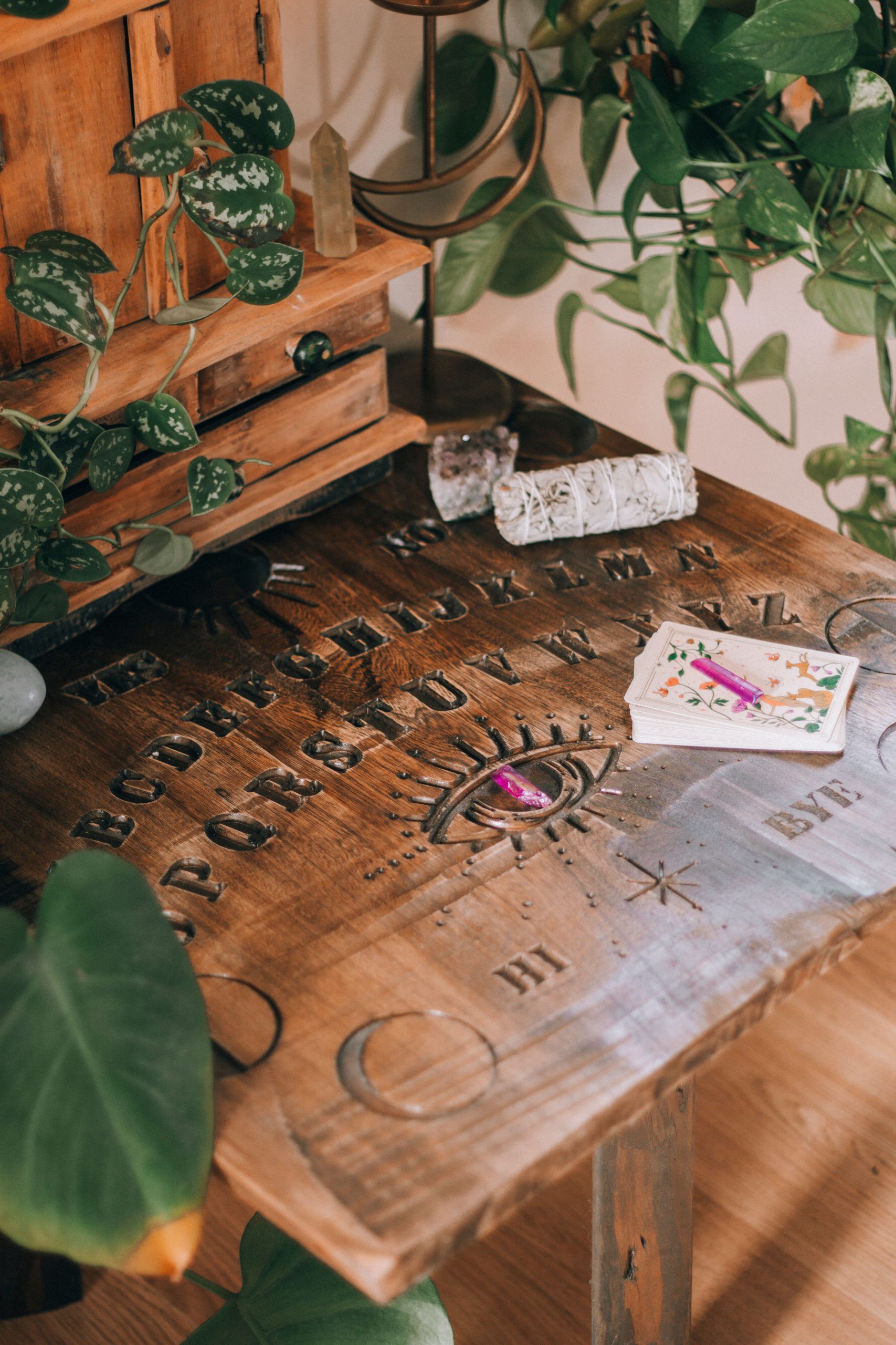 A mystical setup with a Ouija board, crystals, and tarot cards surrounded by greenery.
