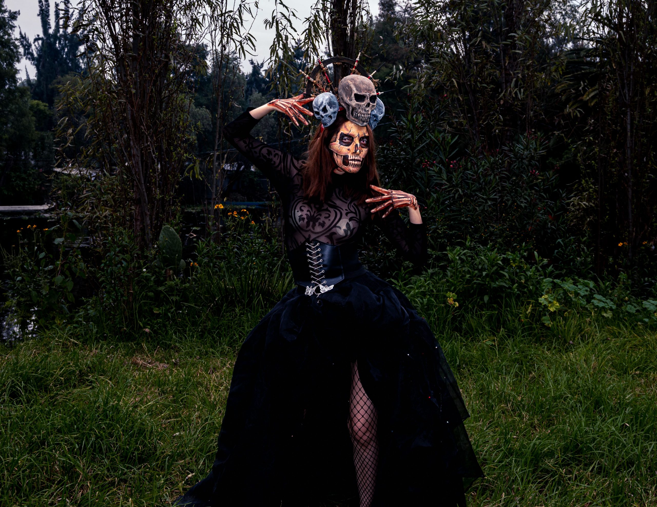 A person in gothic costume and skeleton makeup in a dark outdoor setting, evoking Halloween vibes.