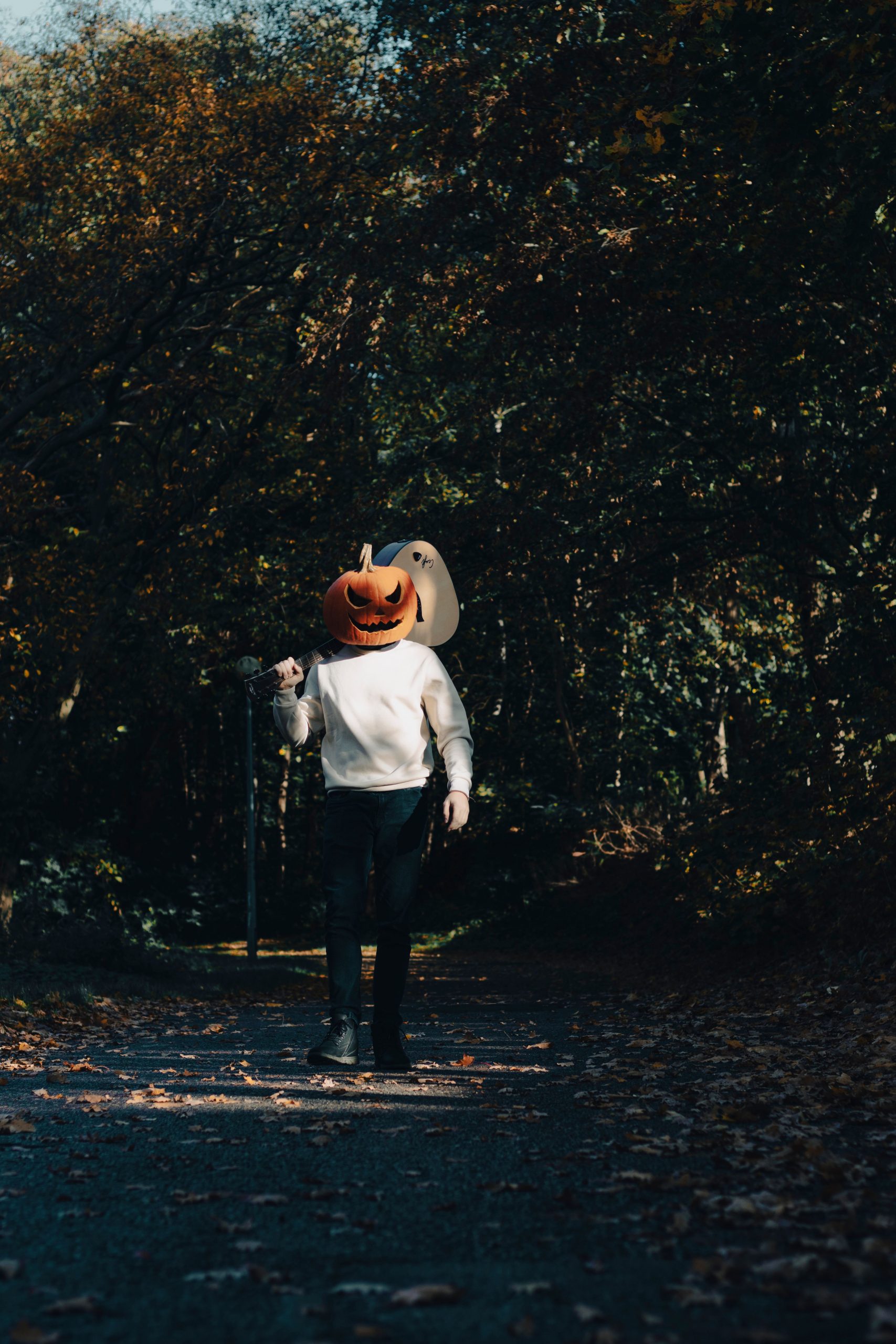 A person with a carved pumpkin head walking in a forest, holding a guitar during the fall season.