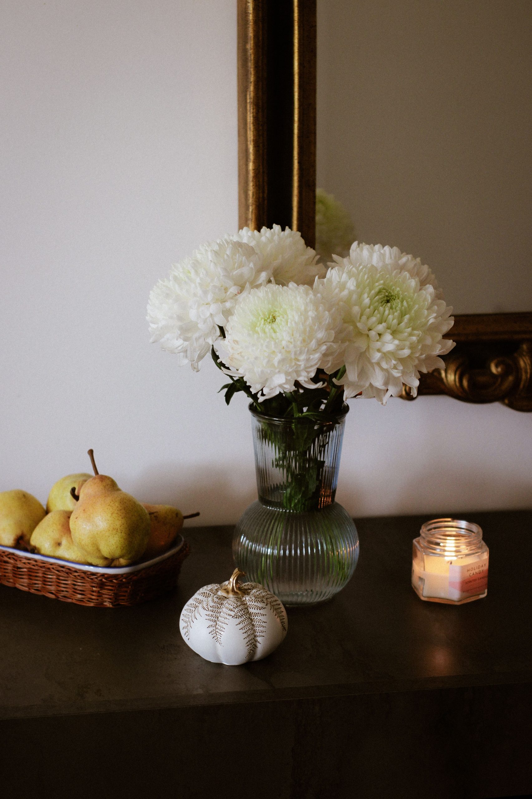 A serene floral setup with white chrysanthemums, pears, and candle on a mantle.