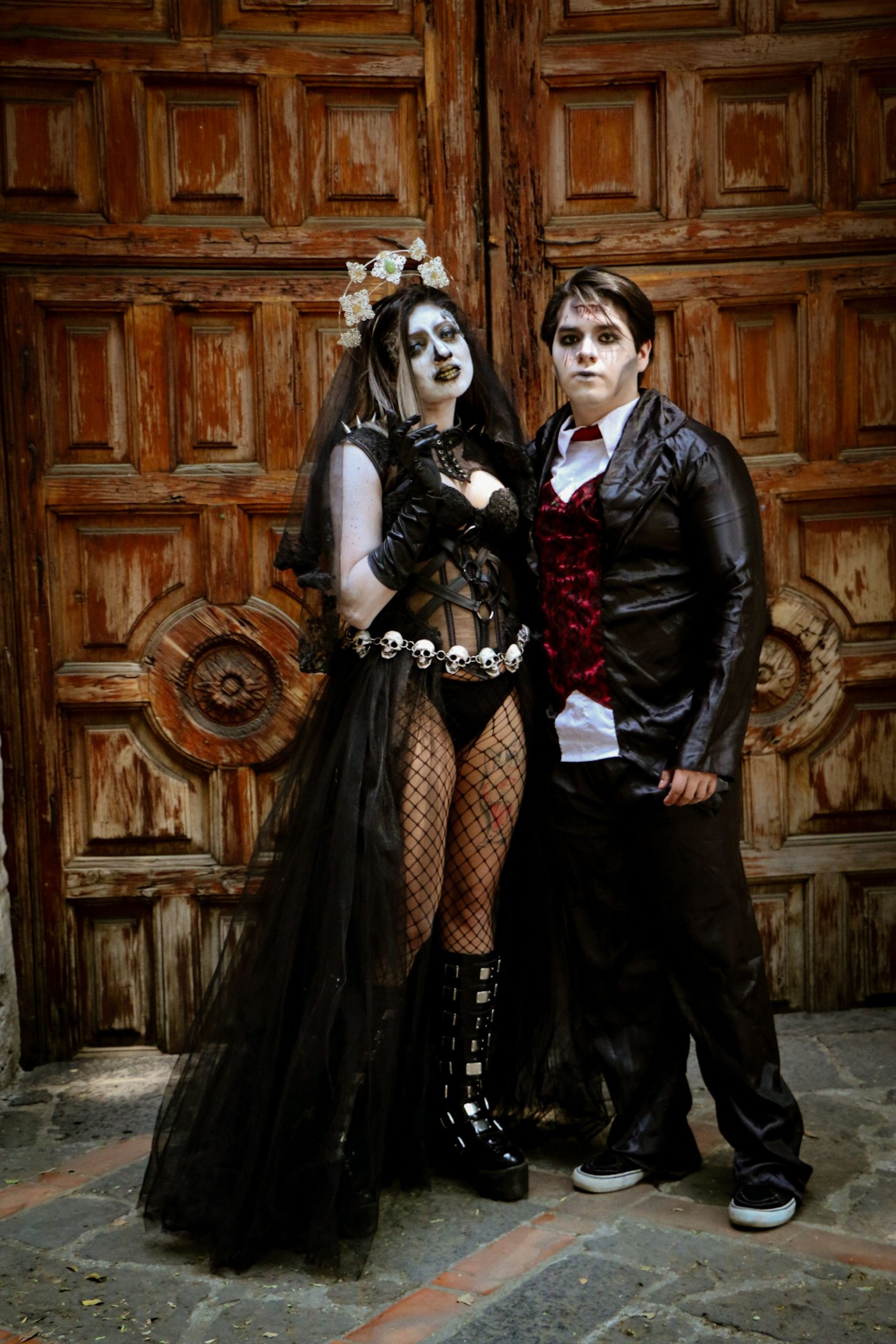 A spooky couple in Gothic Halloween costumes stands before a vintage wooden door.