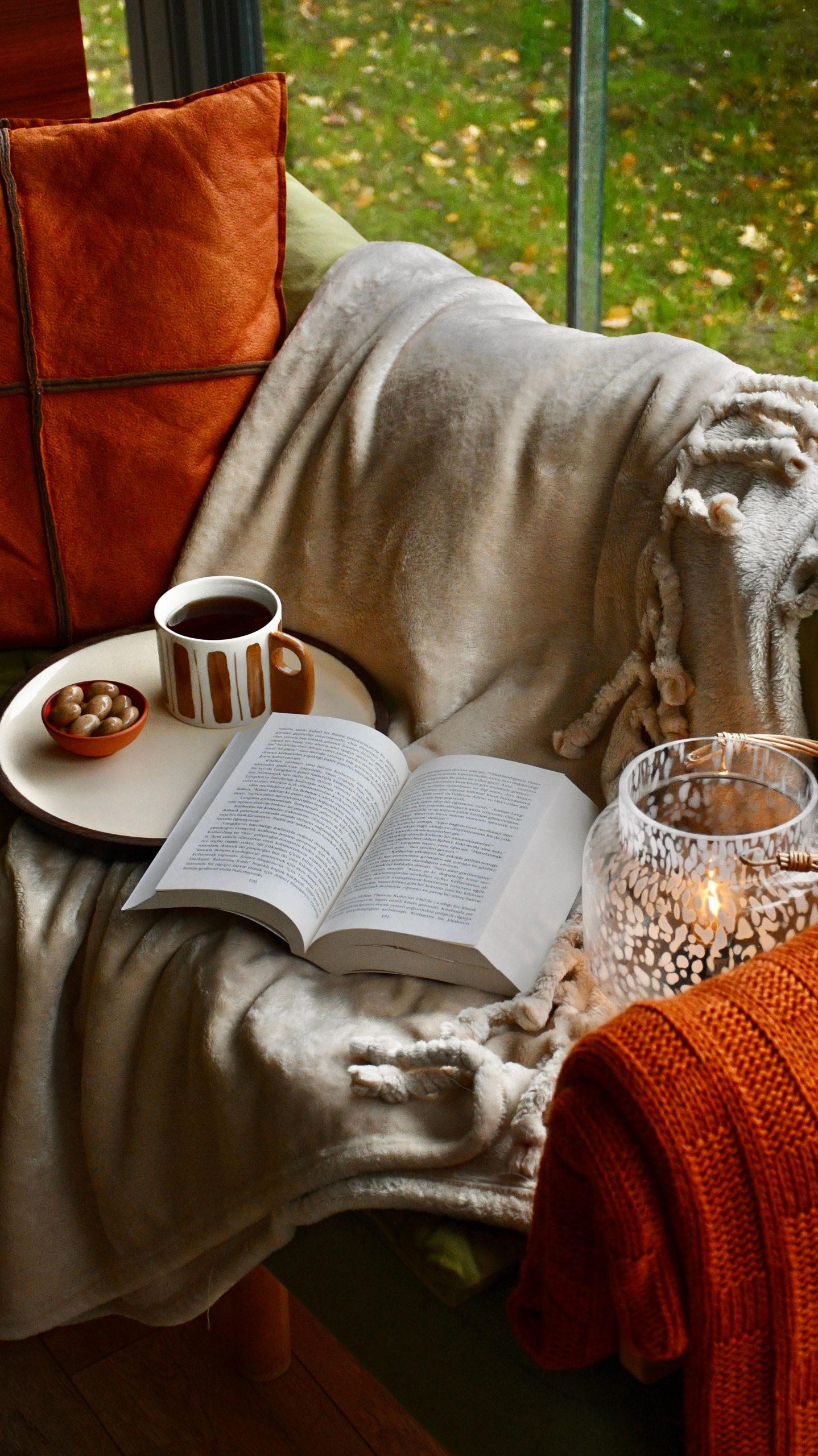 A warm and inviting autumn reading nook featuring a blanket, open book, and mug with a candle and cozy pillows.