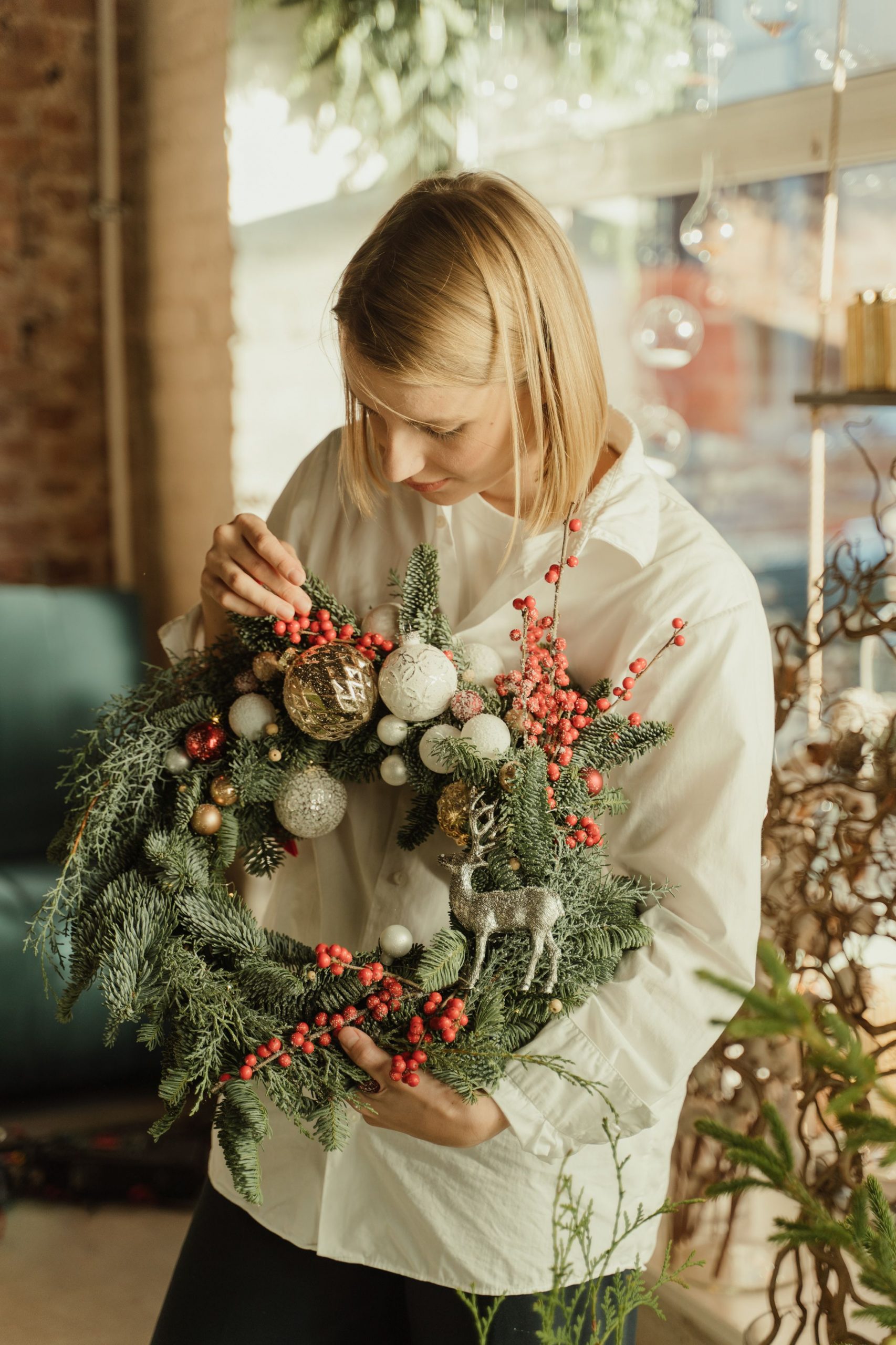 A woman decorates a Christmas wreath with ornaments in a cozy indoor setting.