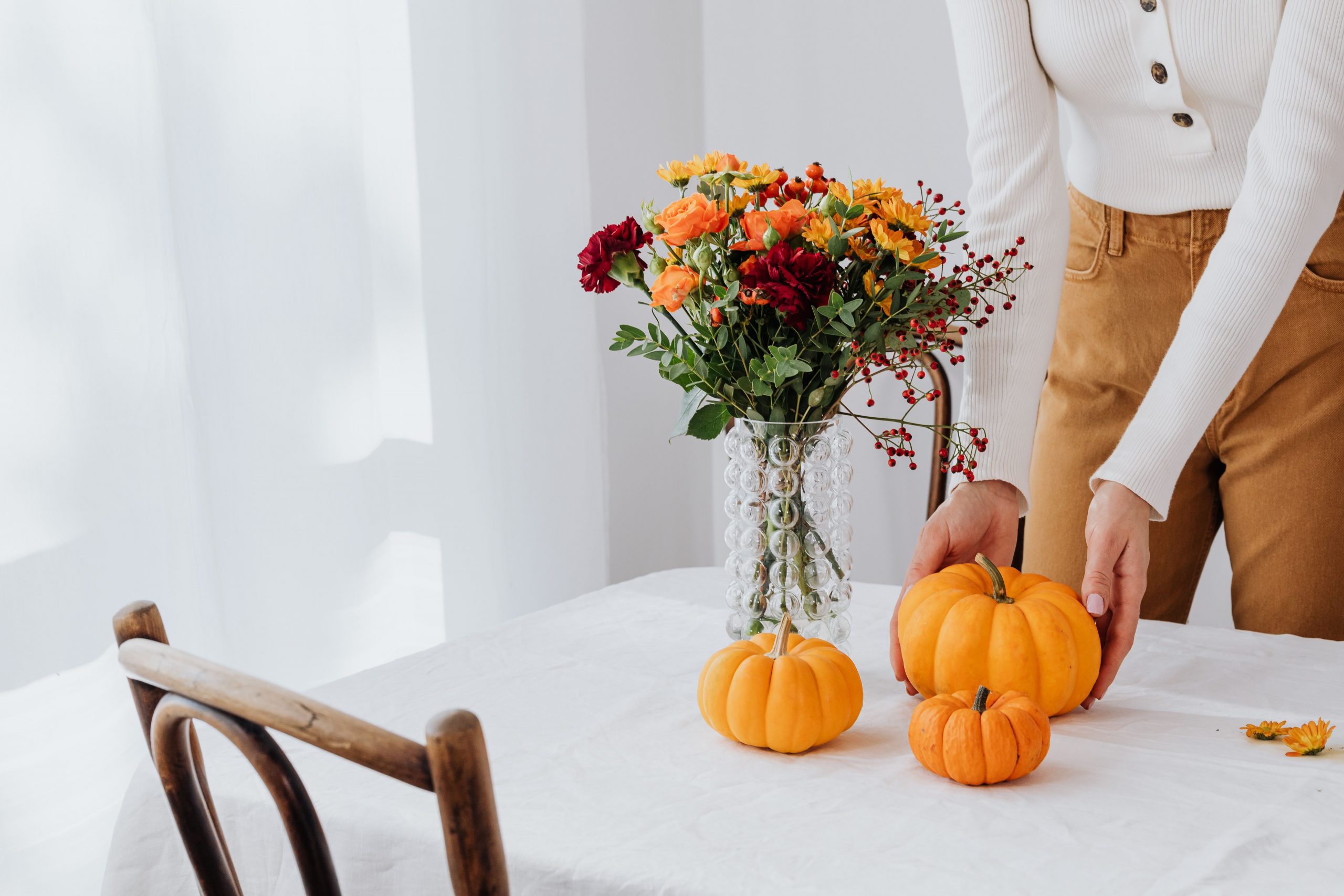 A woman decorates a table with pumpkins and a floral arrangement in a glass vase for fall.