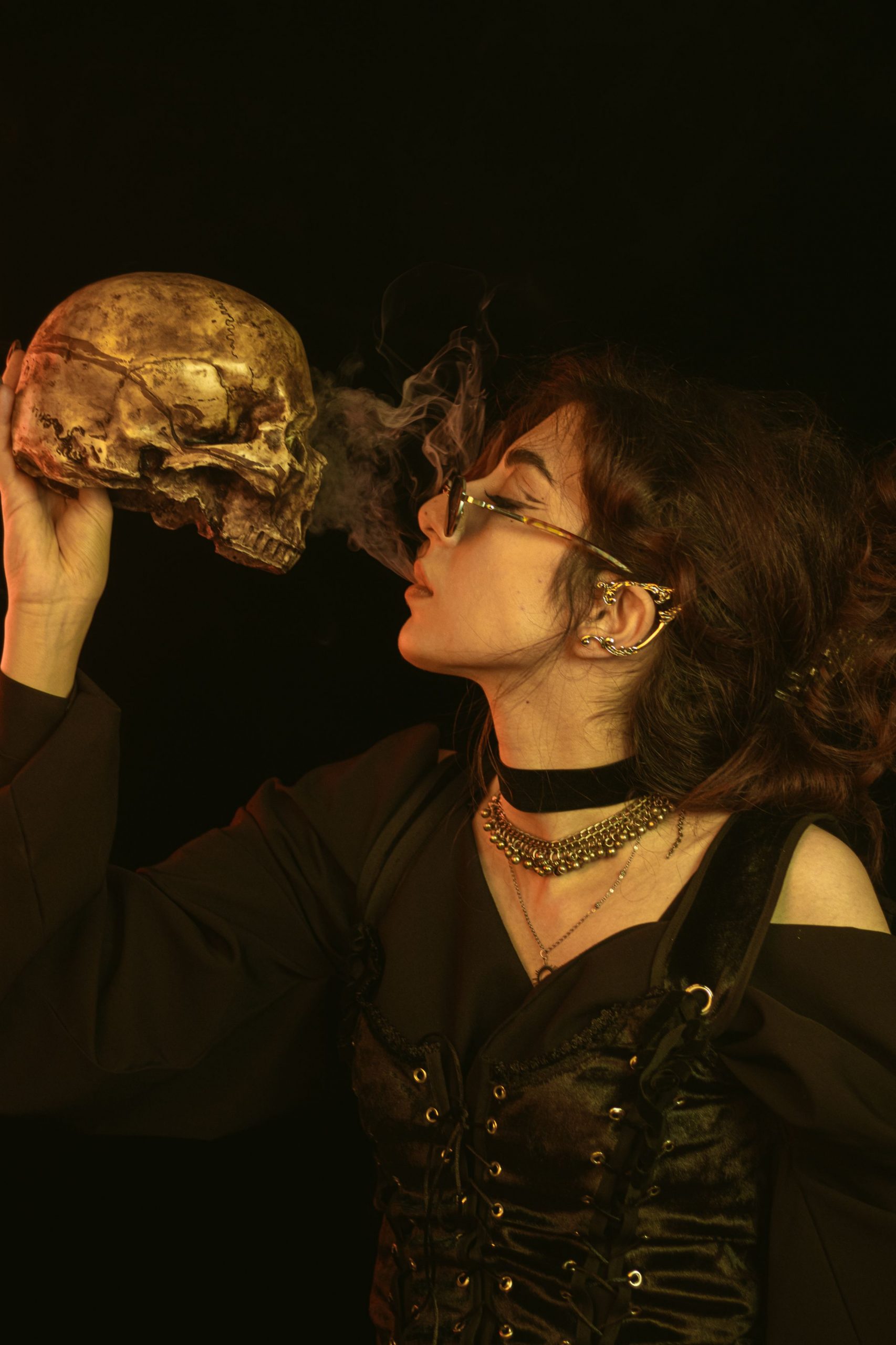 A woman dressed in gothic attire blows smoke onto a skull against a dark backdrop.