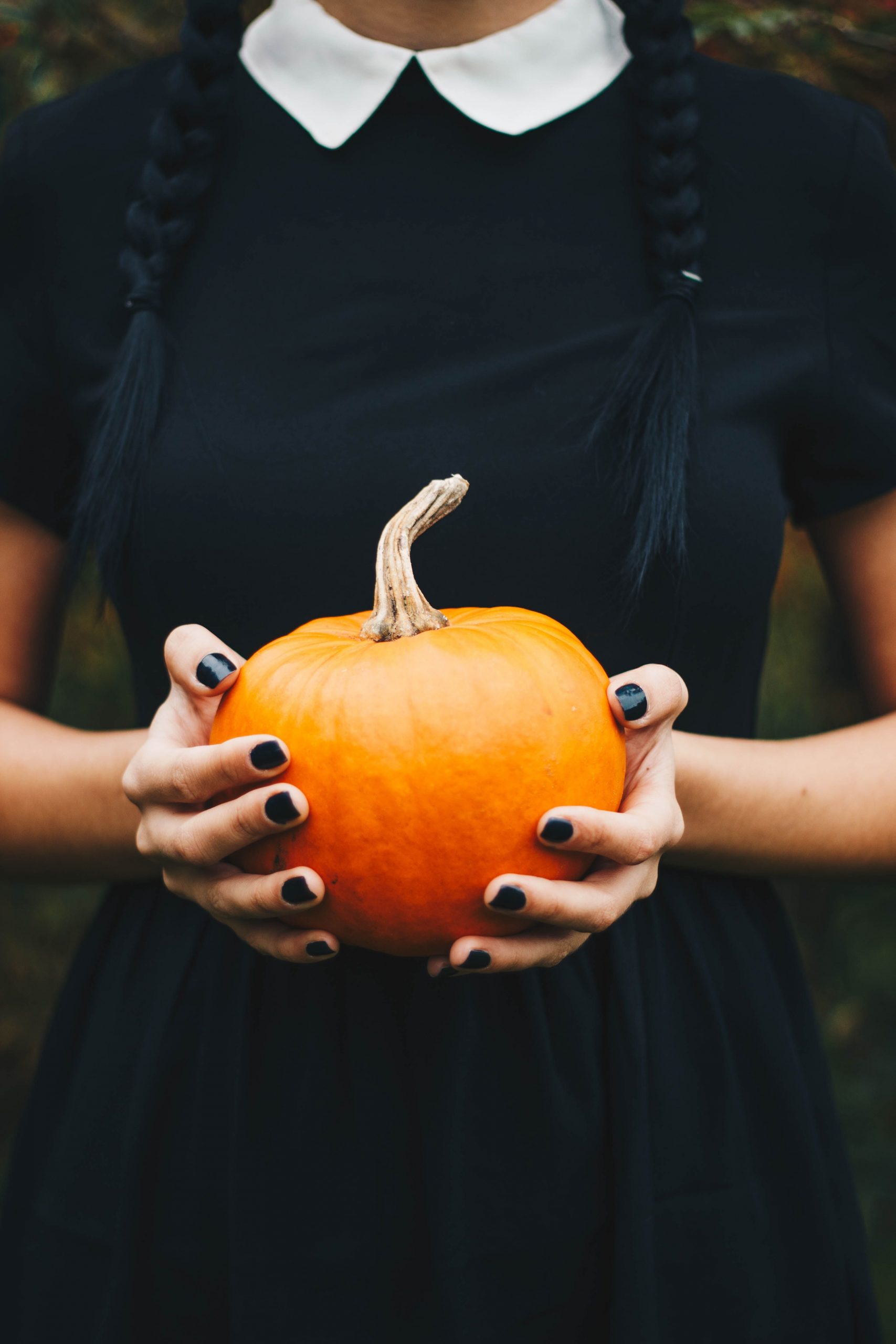 A woman in a black dress with braided hair holding a pumpkin outdoors, perfect for fall and Halloween themes.