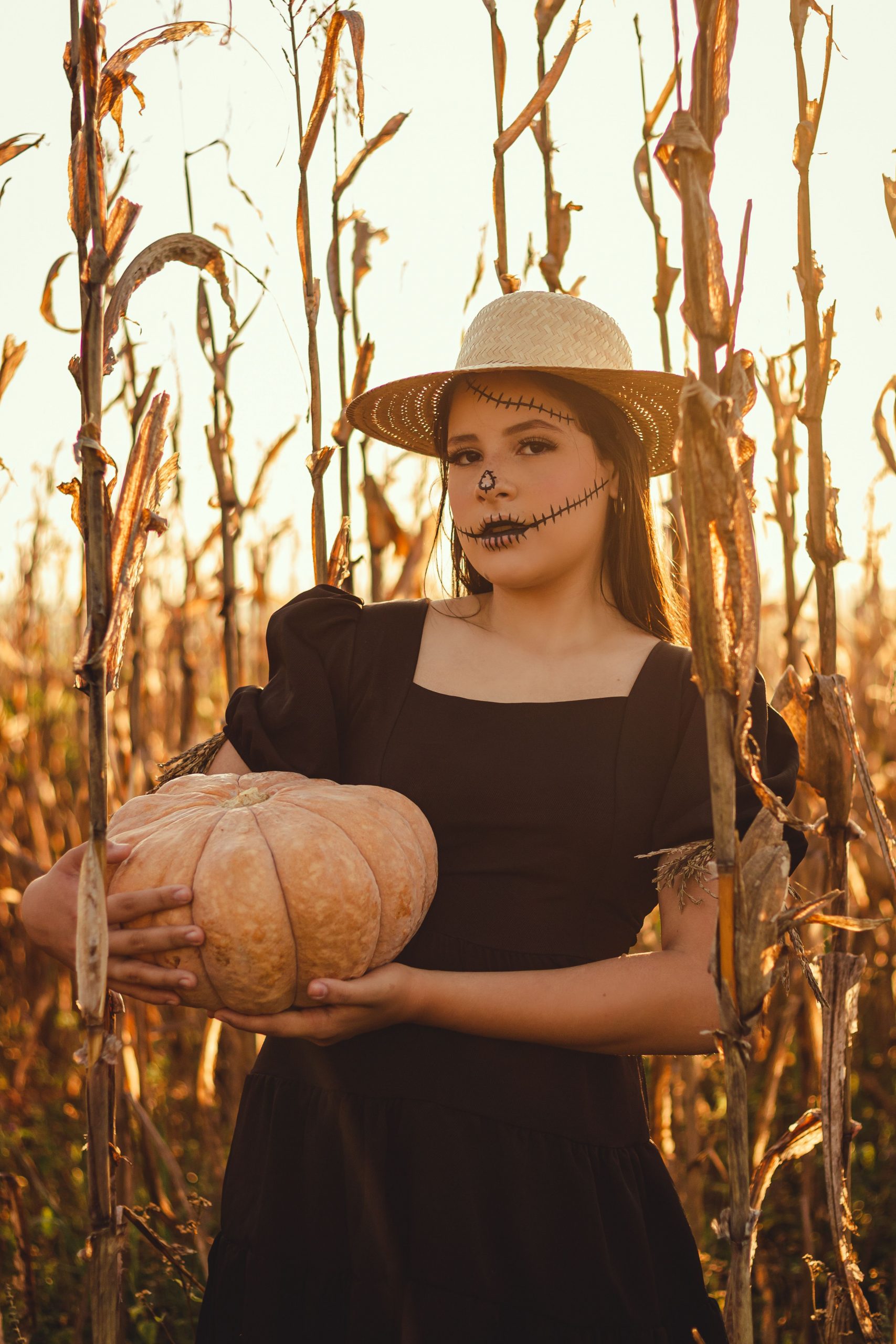 A woman in Halloween makeup and a black dress holds a pumpkin in a sunlit cornfield.