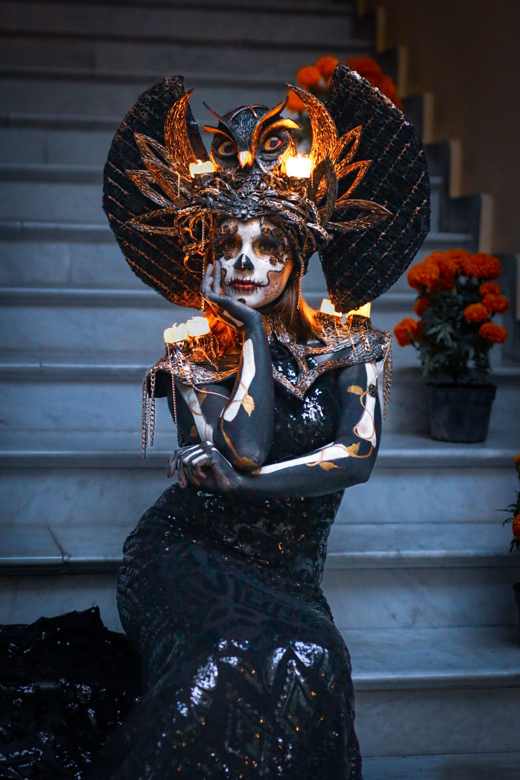 A woman poses in an elaborate Halloween costume with face paint and headpiece on steps, surrounded by candles and flowers.