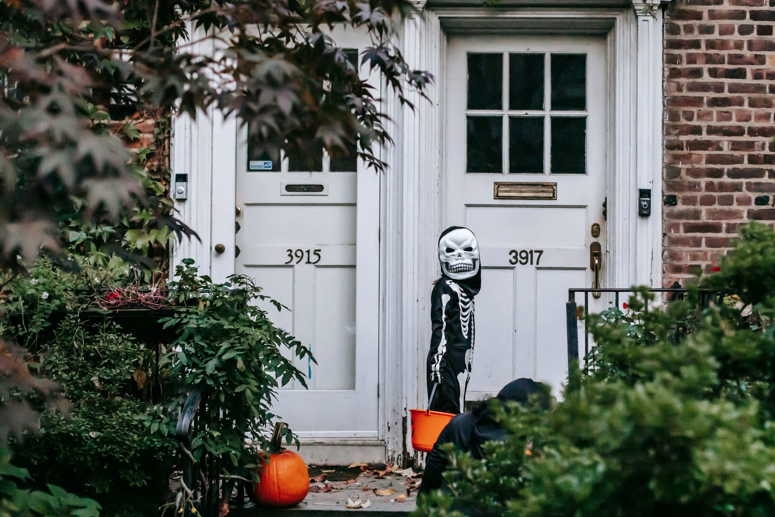 Anonymous child in Halloween costume and mask standing with bucket near house entrance while friend hiding in bushes in daylight
