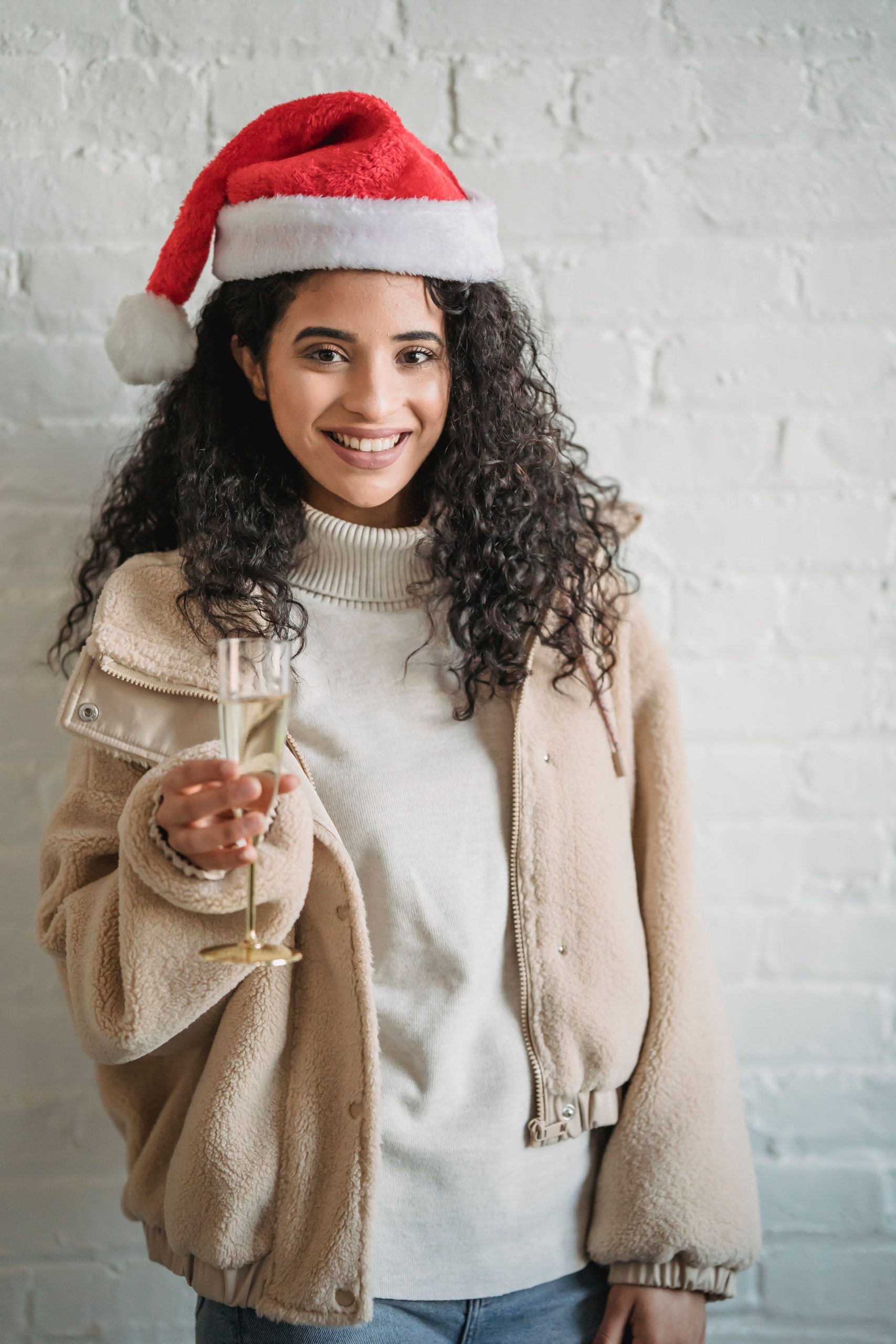 Attractive young ethnic female in warm sweater and red Santa hat standing with glass of sparkling champagne and looking at camera with charming smile