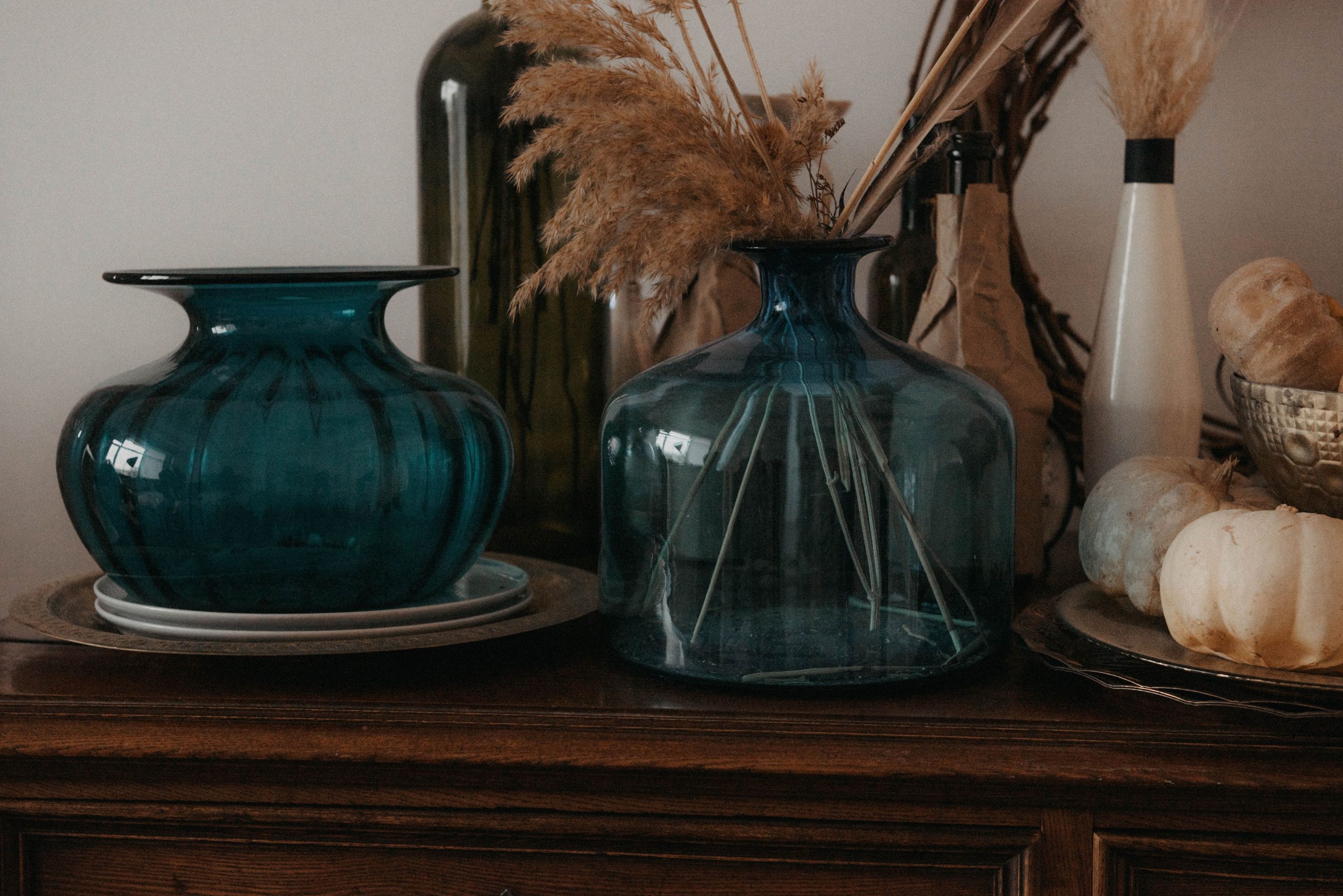Autumn-themed arrangement with glass vases, dried plants, and pumpkins on a wooden dresser.