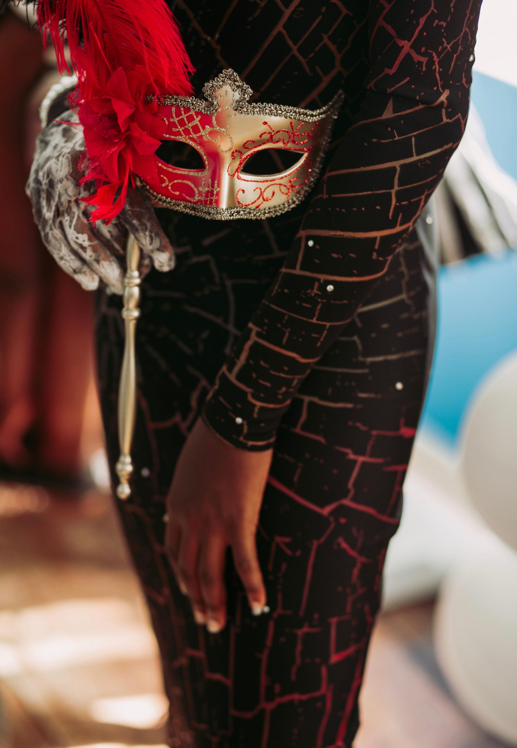 Close-up of an elegant masquerade mask with red feathers held by a woman in a stylish black dress, perfect for parties.