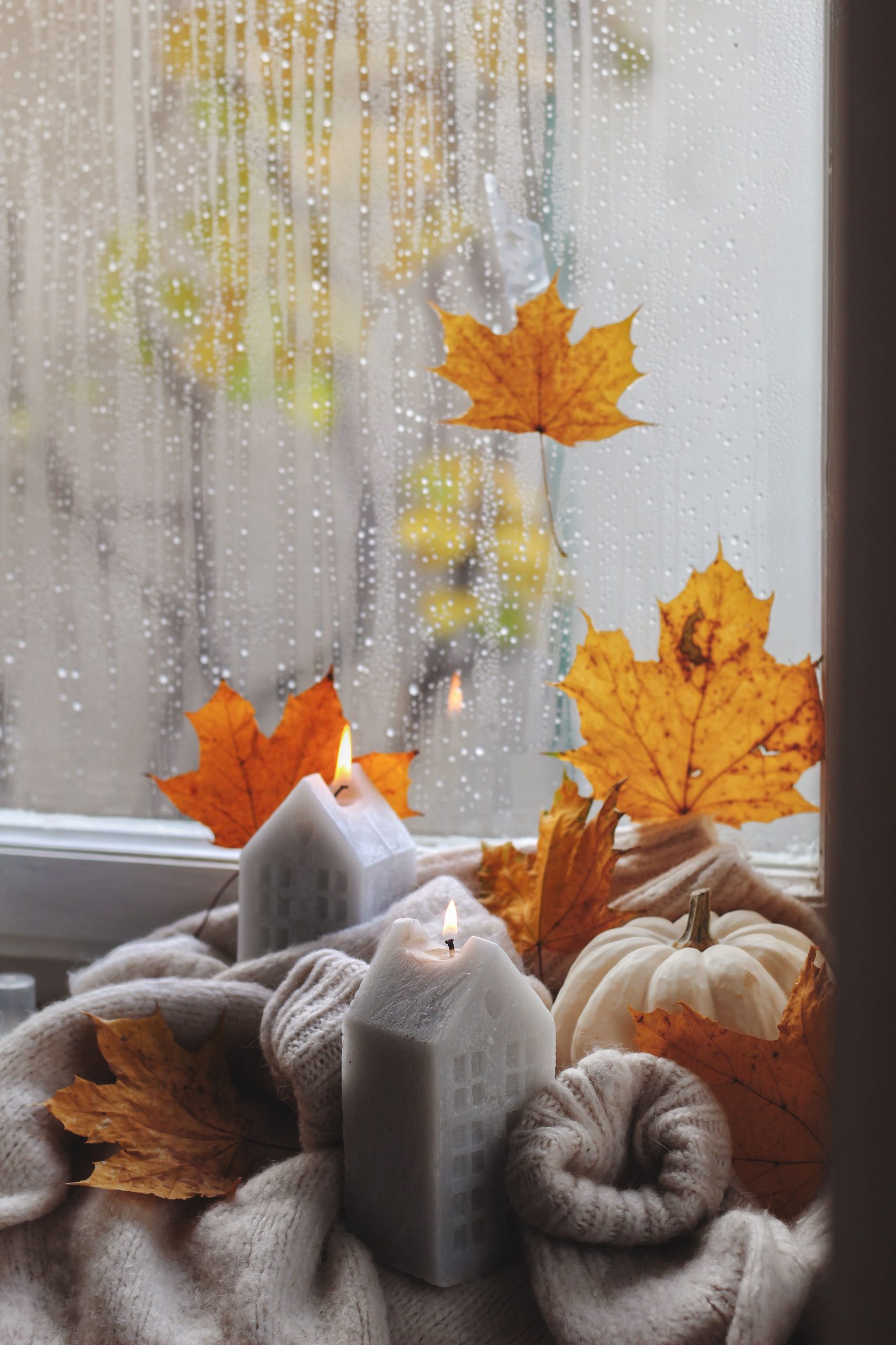 Comforting fall scene with candles, sweater, leaves, and pumpkin on windowsill.