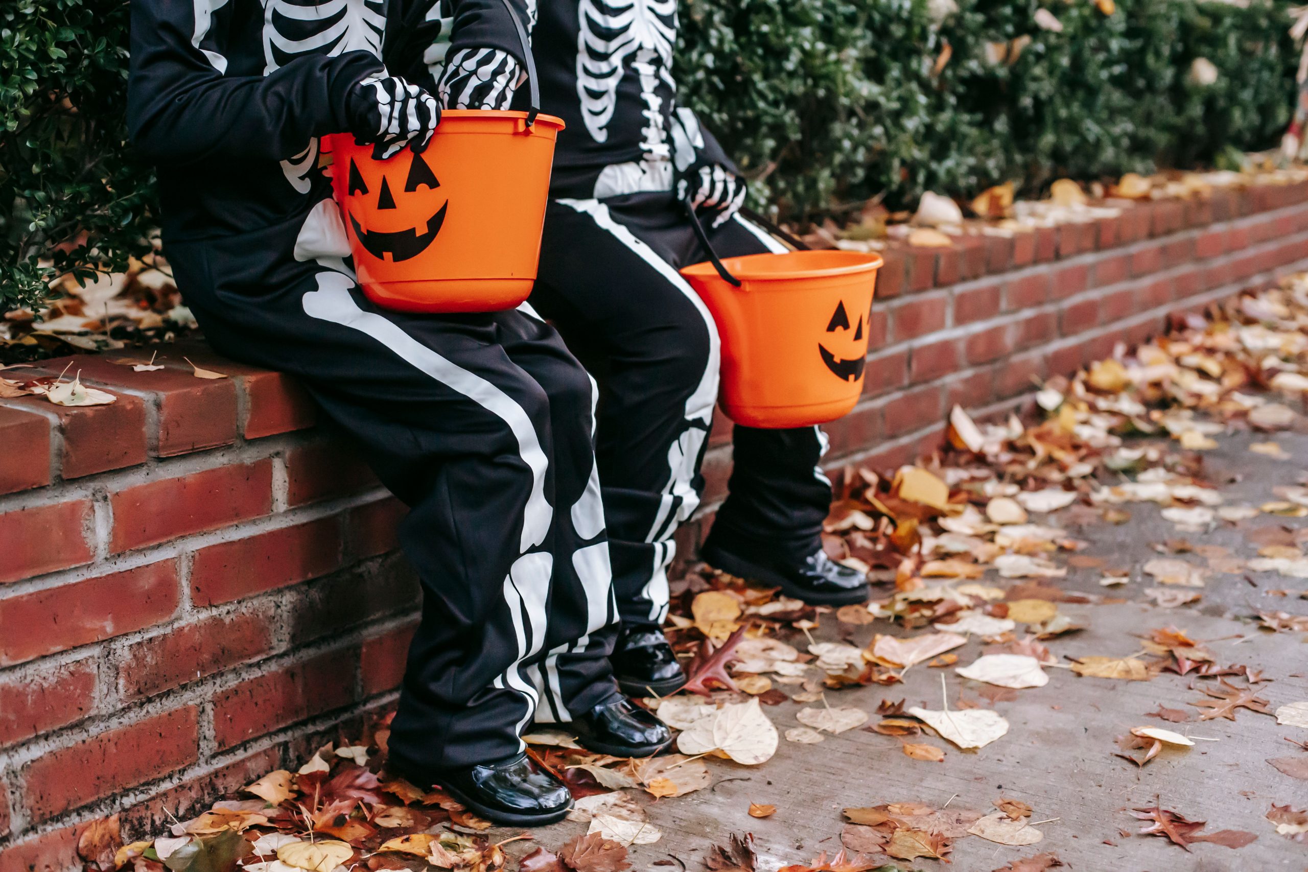 Crop anonymous children dressed as scary skeletons sitting on red brick fence with jack o lantern buckets on Halloween day