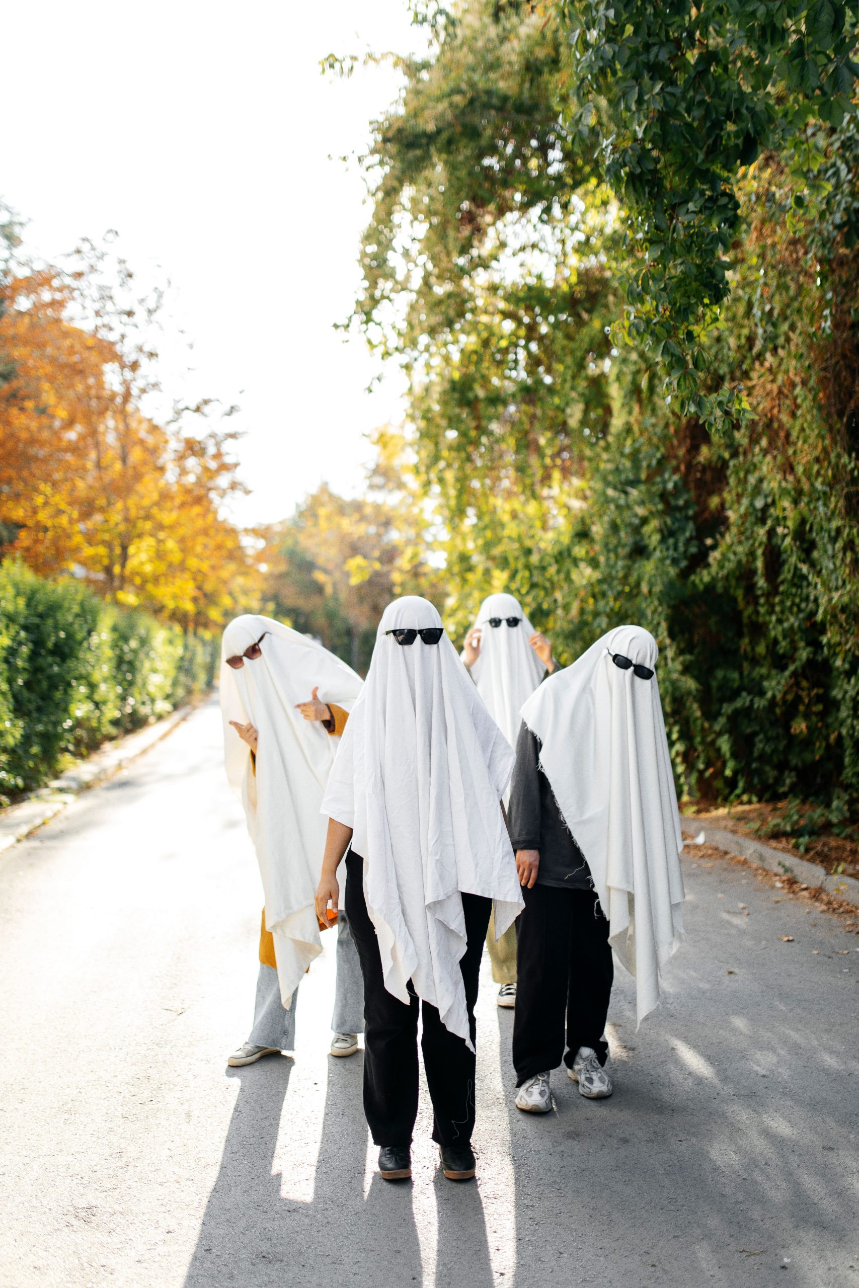 Four people dressed as ghosts with sunglasses enjoying a sunny autumn day on a city street.