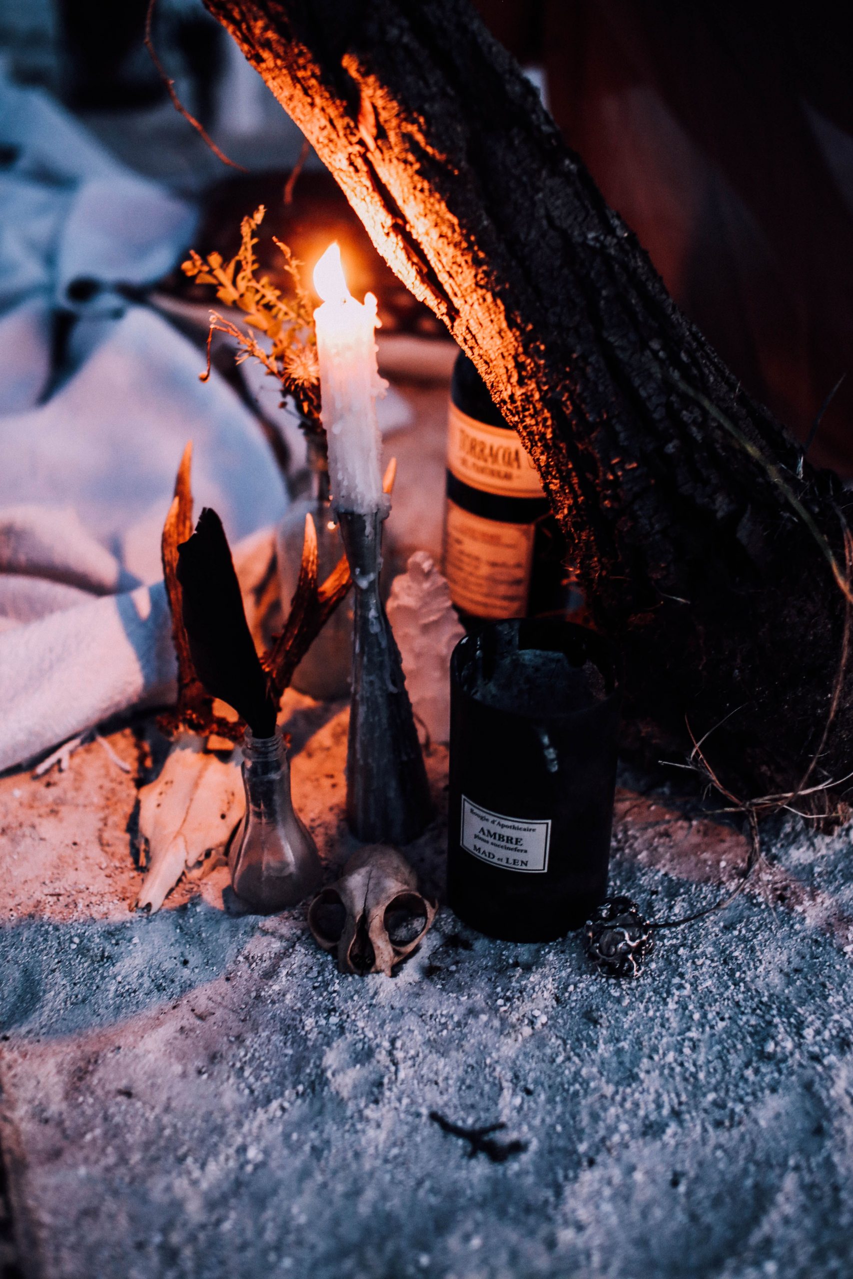 From above of gloomy composition with bottle of alcohol and skull near burning candle and bottles placed on white sand near peace of wood