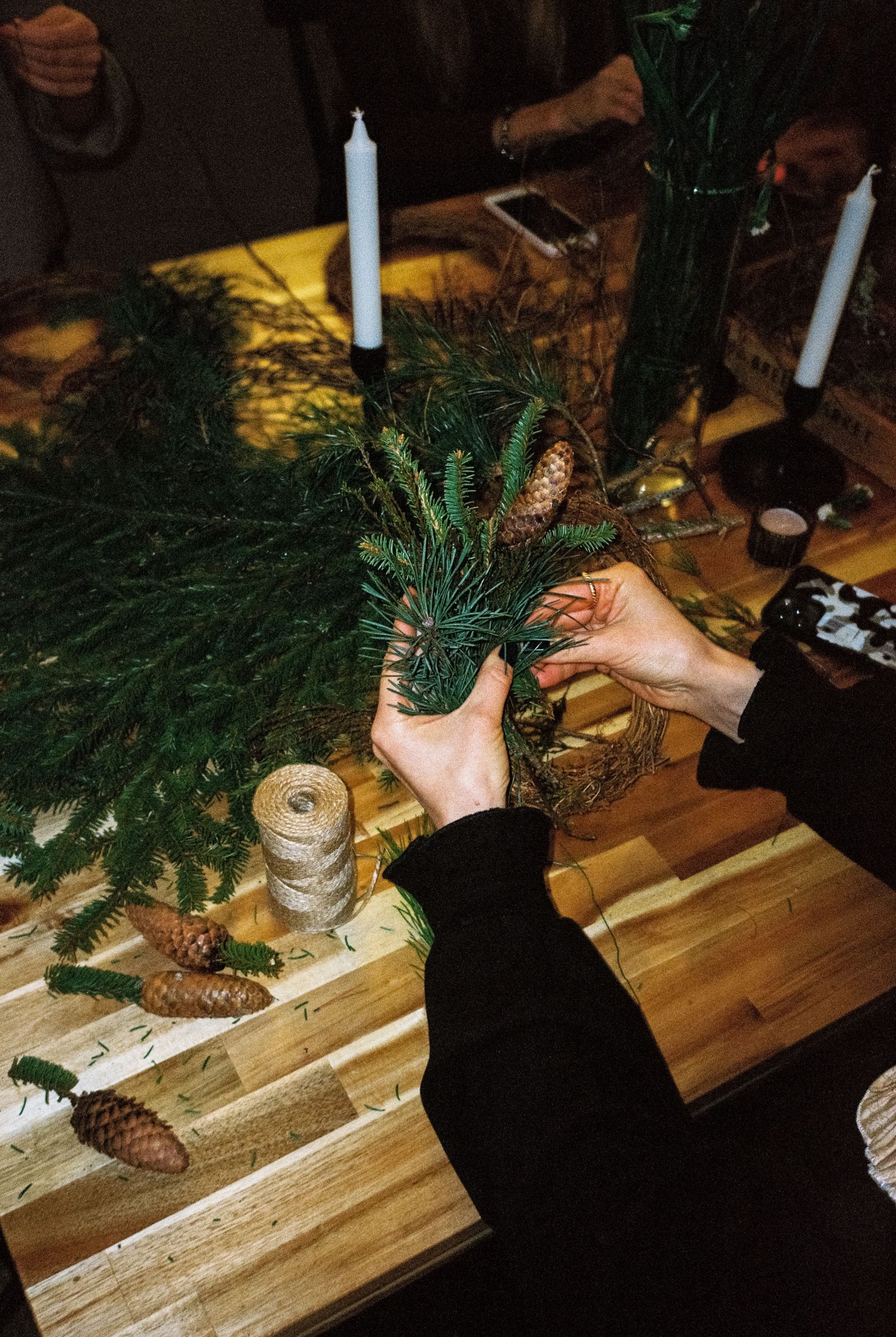 Hands craft a Christmas wreath with pine branches and candles on a wooden table.