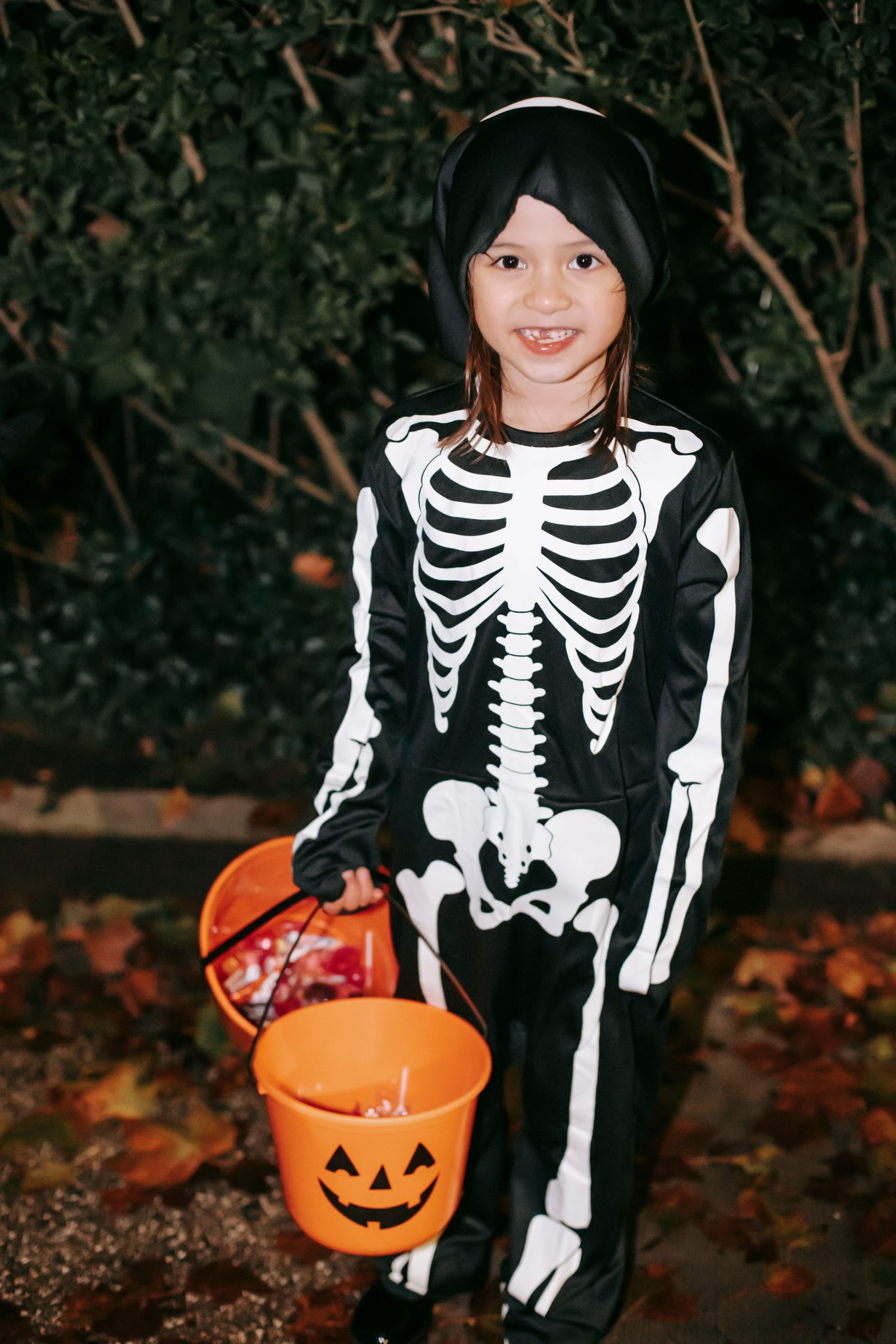Happy girl in skeleton costume and headband with orange pumpkin buckets with sweets in hand on autumn street at night