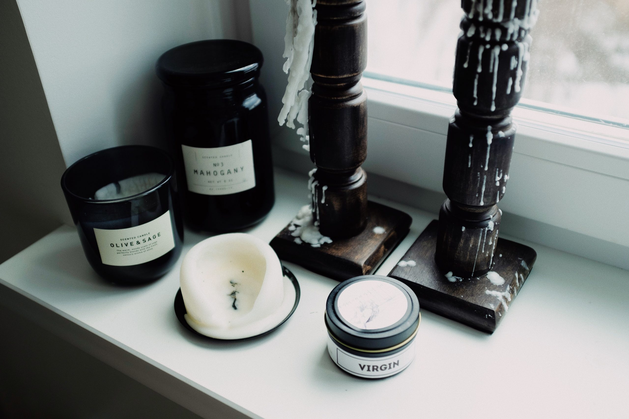 High angle of various candles placed in black glass jars and wooden candlestick arranged on white surface of windowsill in light room