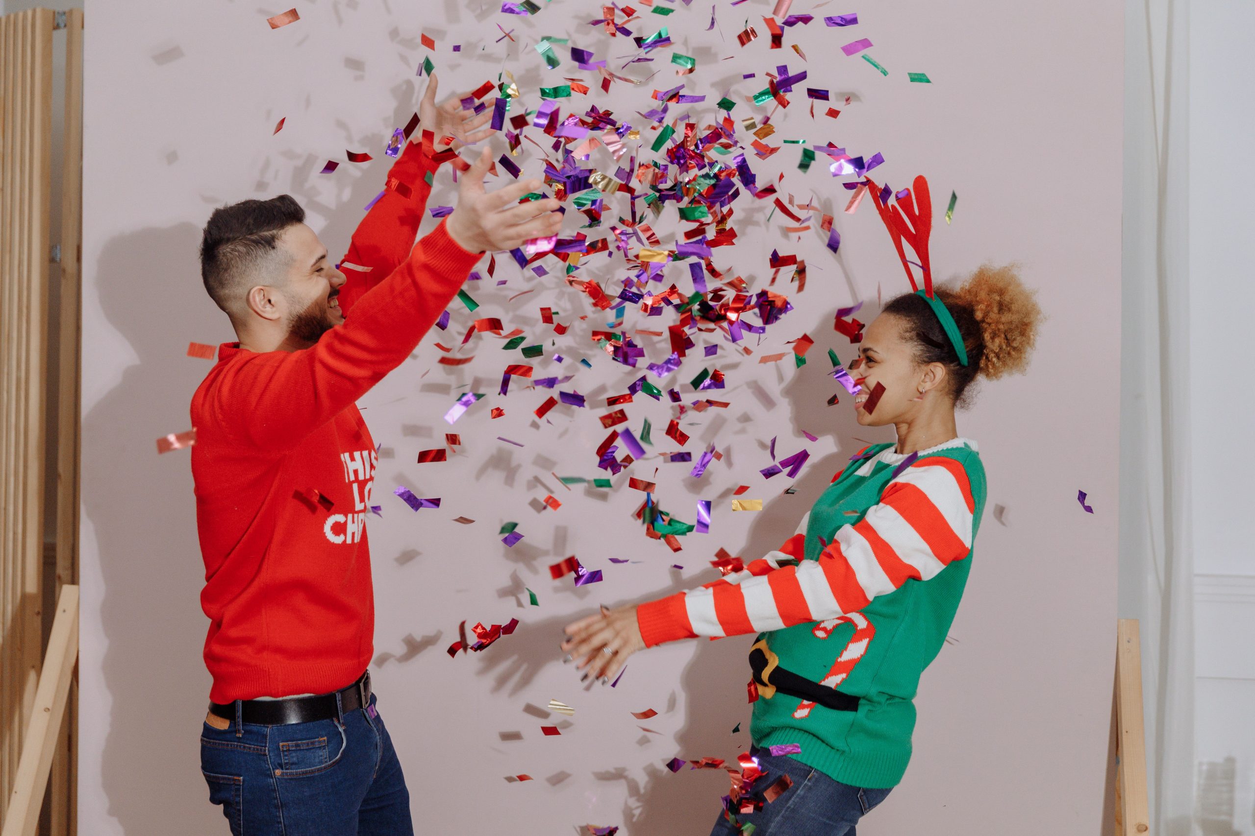 Interracial couple celebrating holidays with colorful confetti in festive sweaters indoors.
