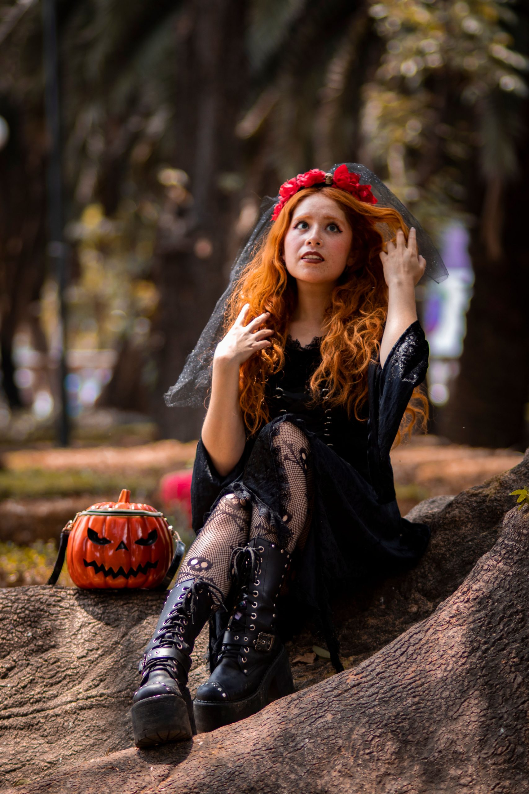 Red-haired woman in black Halloween outfit with pumpkin bag sitting on a rock.