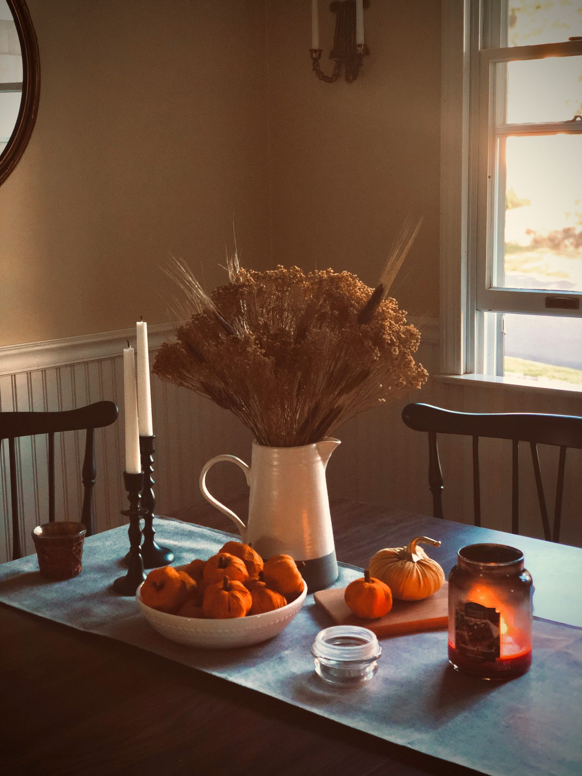 Some wheat grass and other dried fall foliage in a ceramic pitcher vase surrounded by taper candles, a bowl of tiny pumpkins and a lit Yankee candle. It is fall in New England!