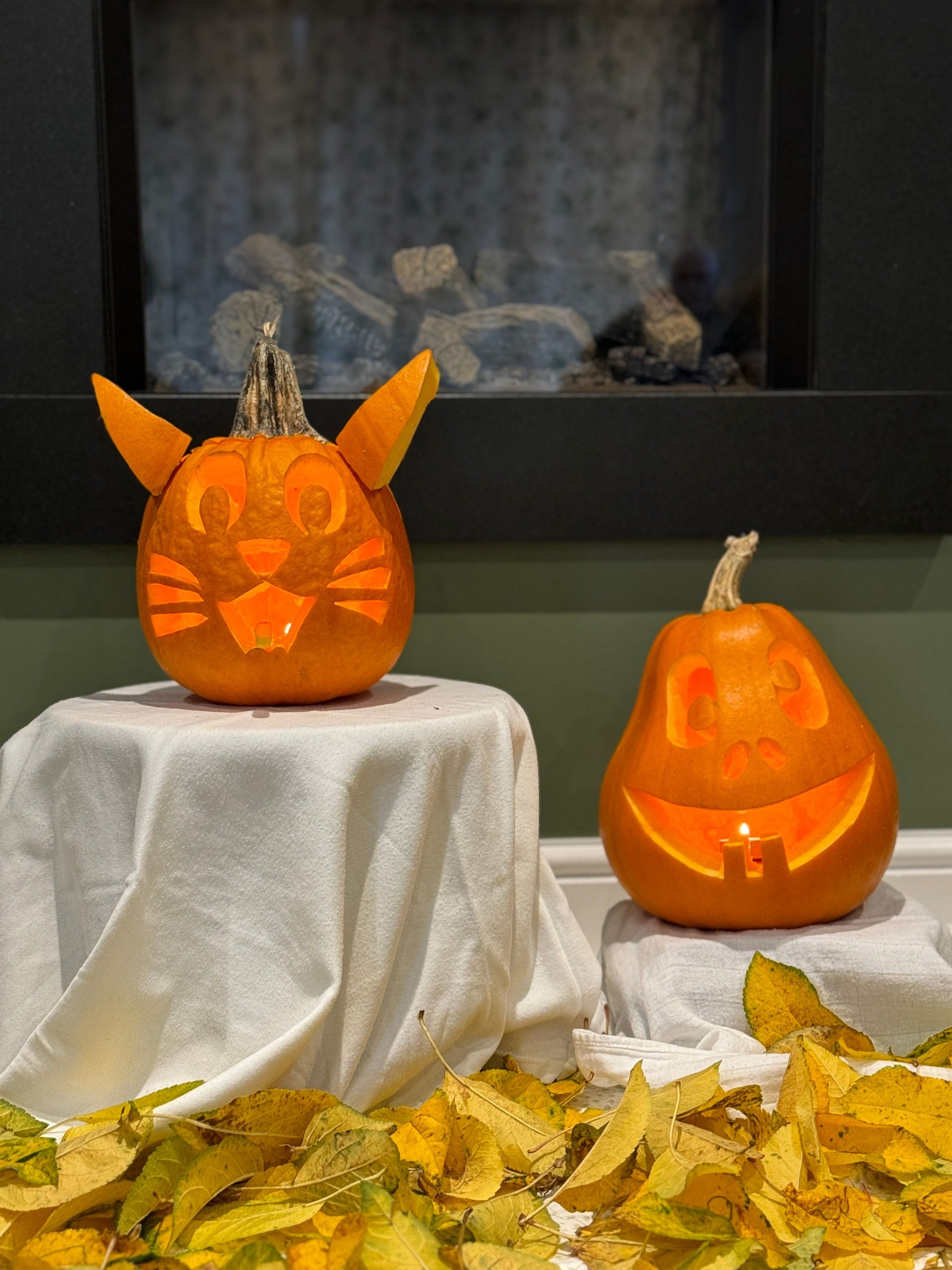 Two uniquely carved pumpkins with autumn leaves in a cozy indoor setting.