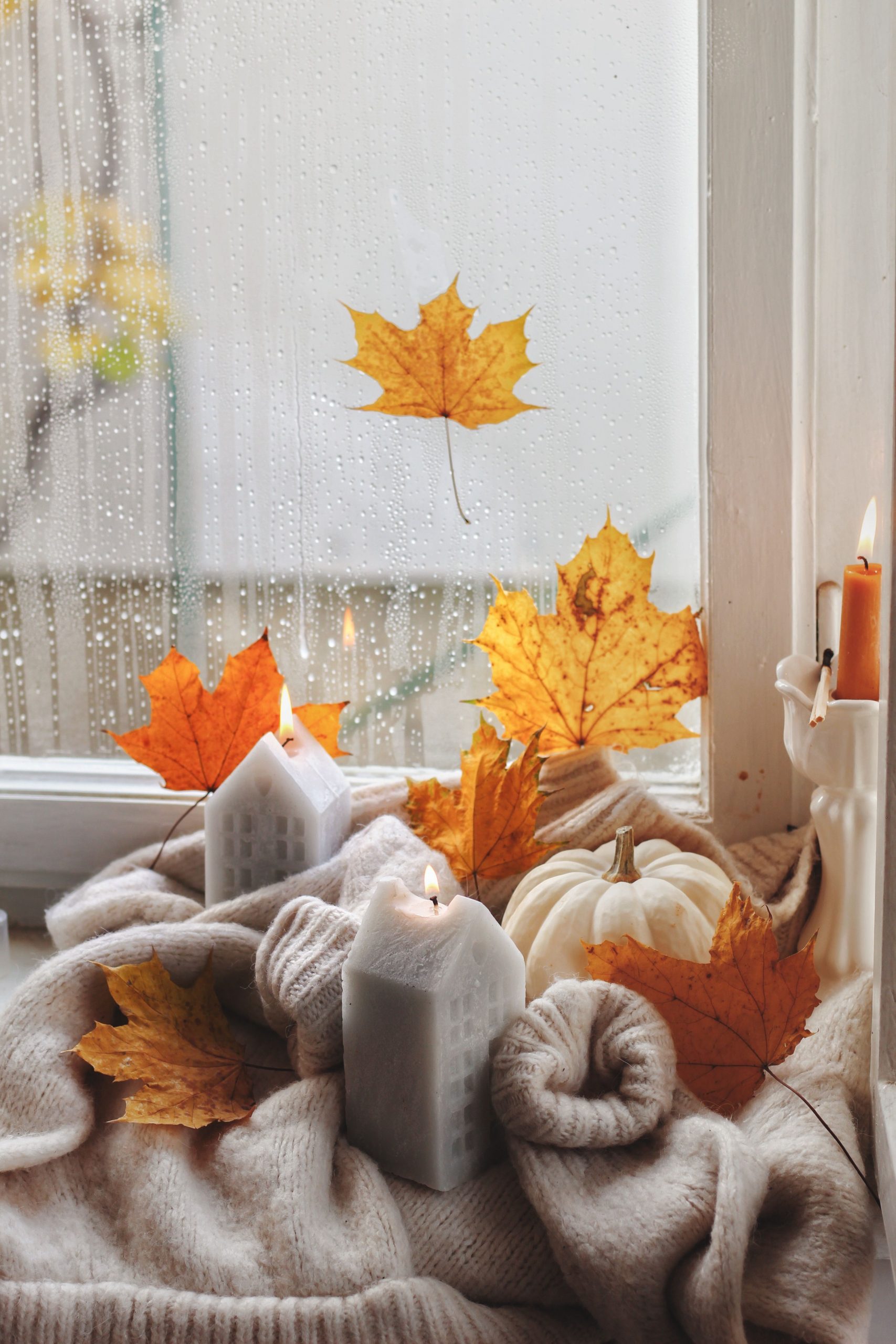 Warm and cozy fall setting featuring candles, a sweater, and autumn leaves by a rainy window.