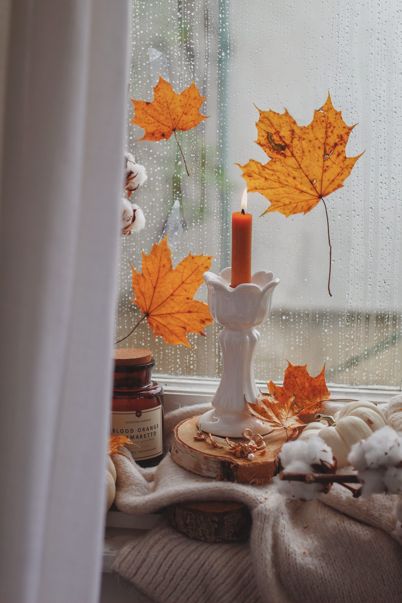 Warm autumn scene with a lit candle by a rain-streaked window adorned with vibrant maple leaves.