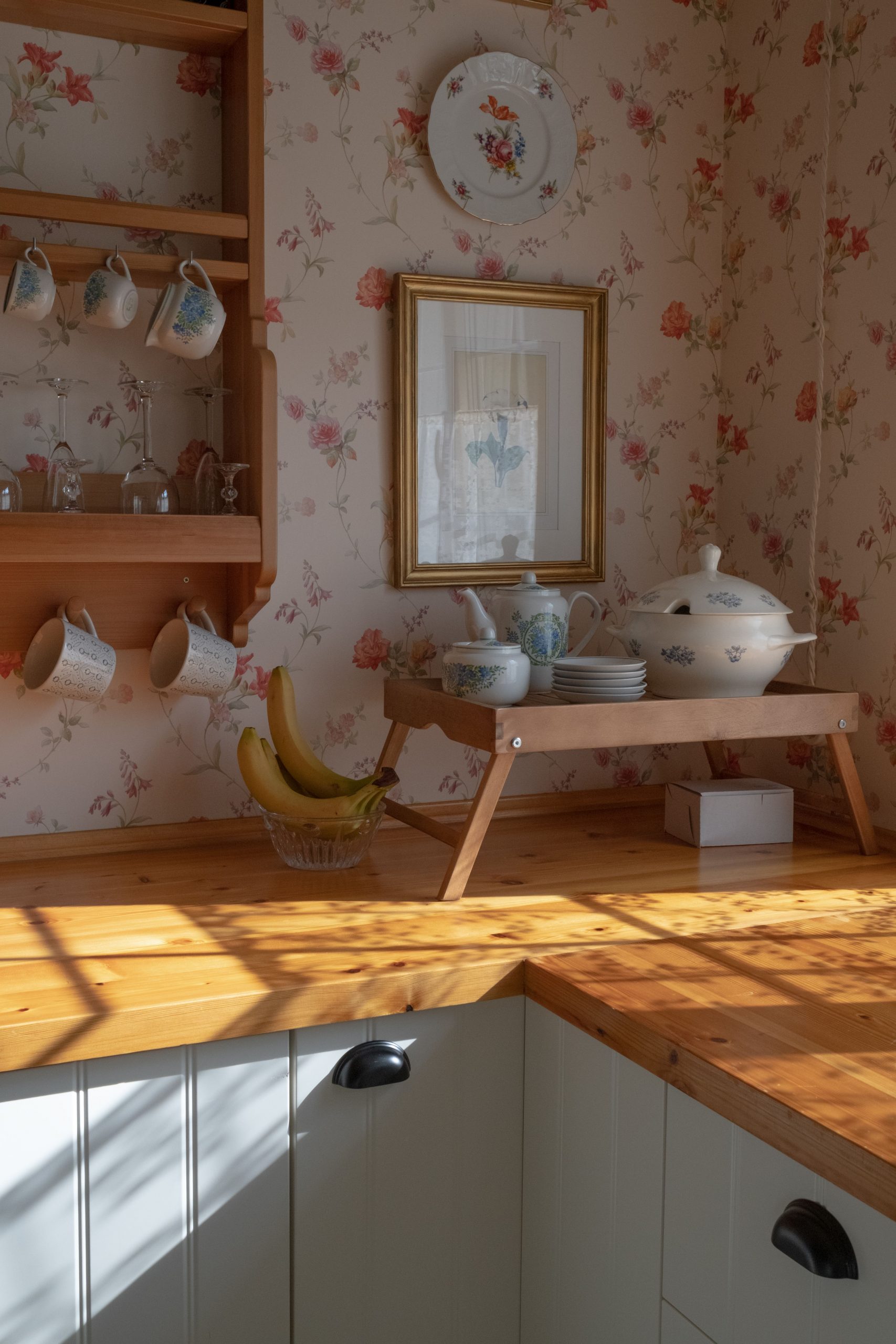 Warm rustic kitchen corner with vintage porcelain and floral wallpaper, captured in sunlight.