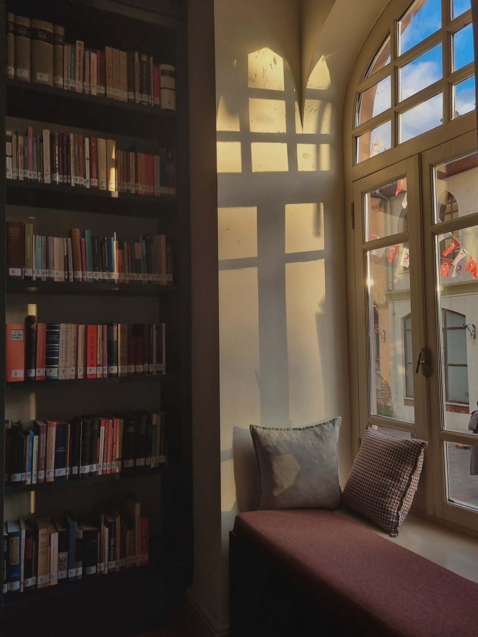 Warm sunlight casts shadows in a library corner with books and cushioned seat by the window.