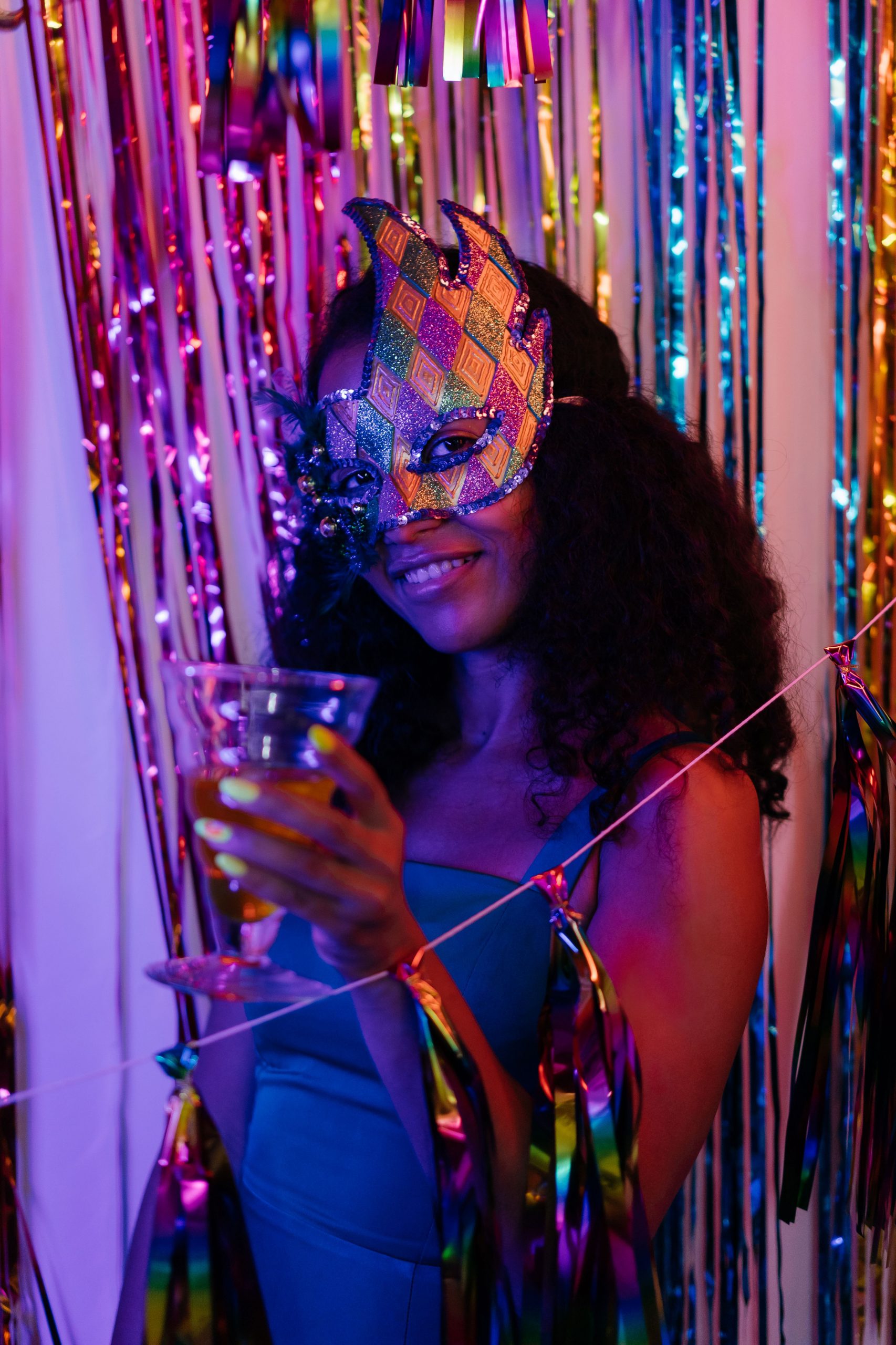 Woman in a vibrant masquerade setting holding a cocktail, with colorful decorations.