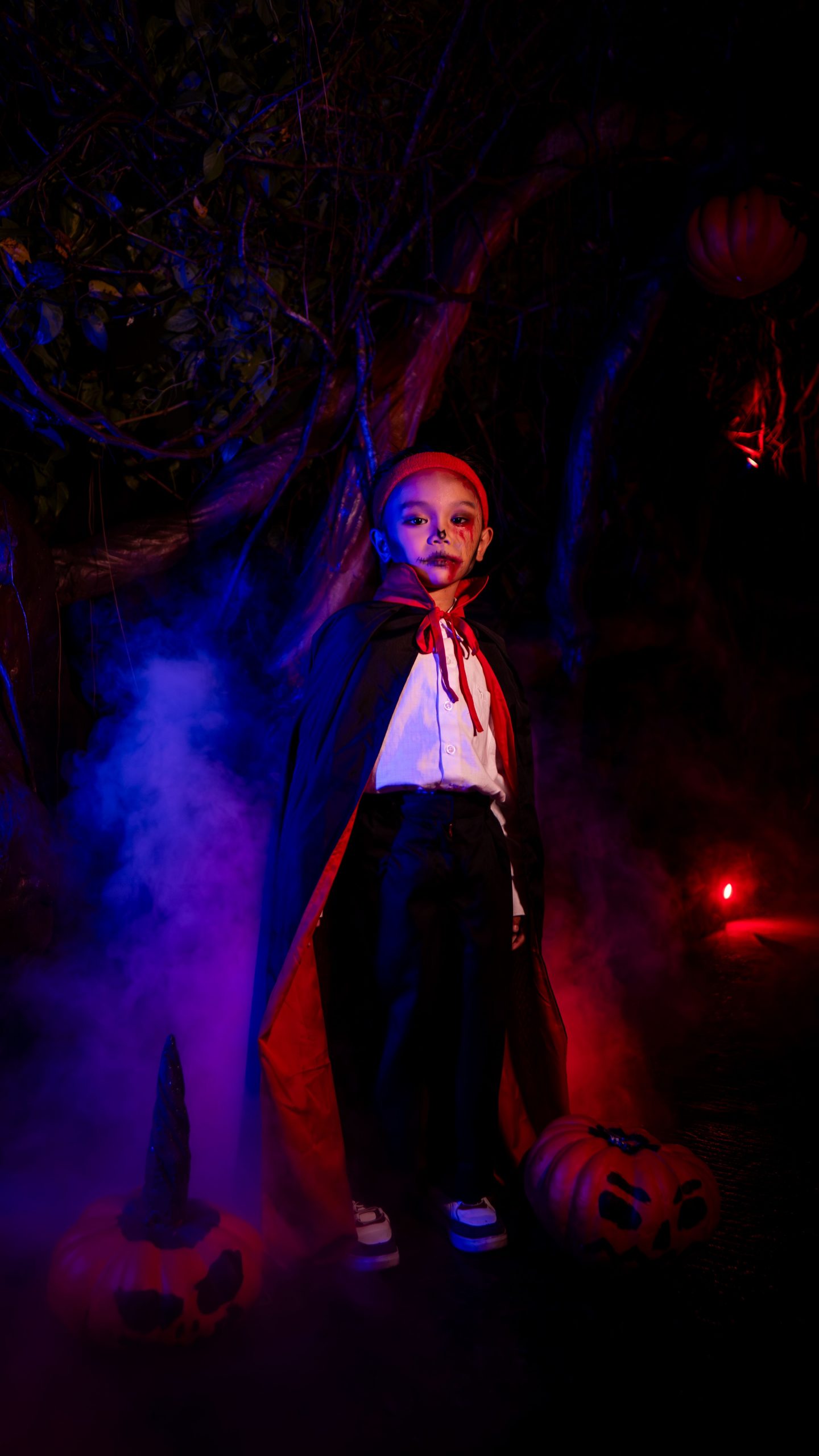 Young child dressed as a vampire, surrounded by eerie fog and Halloween pumpkins in a dark, spooky setting.