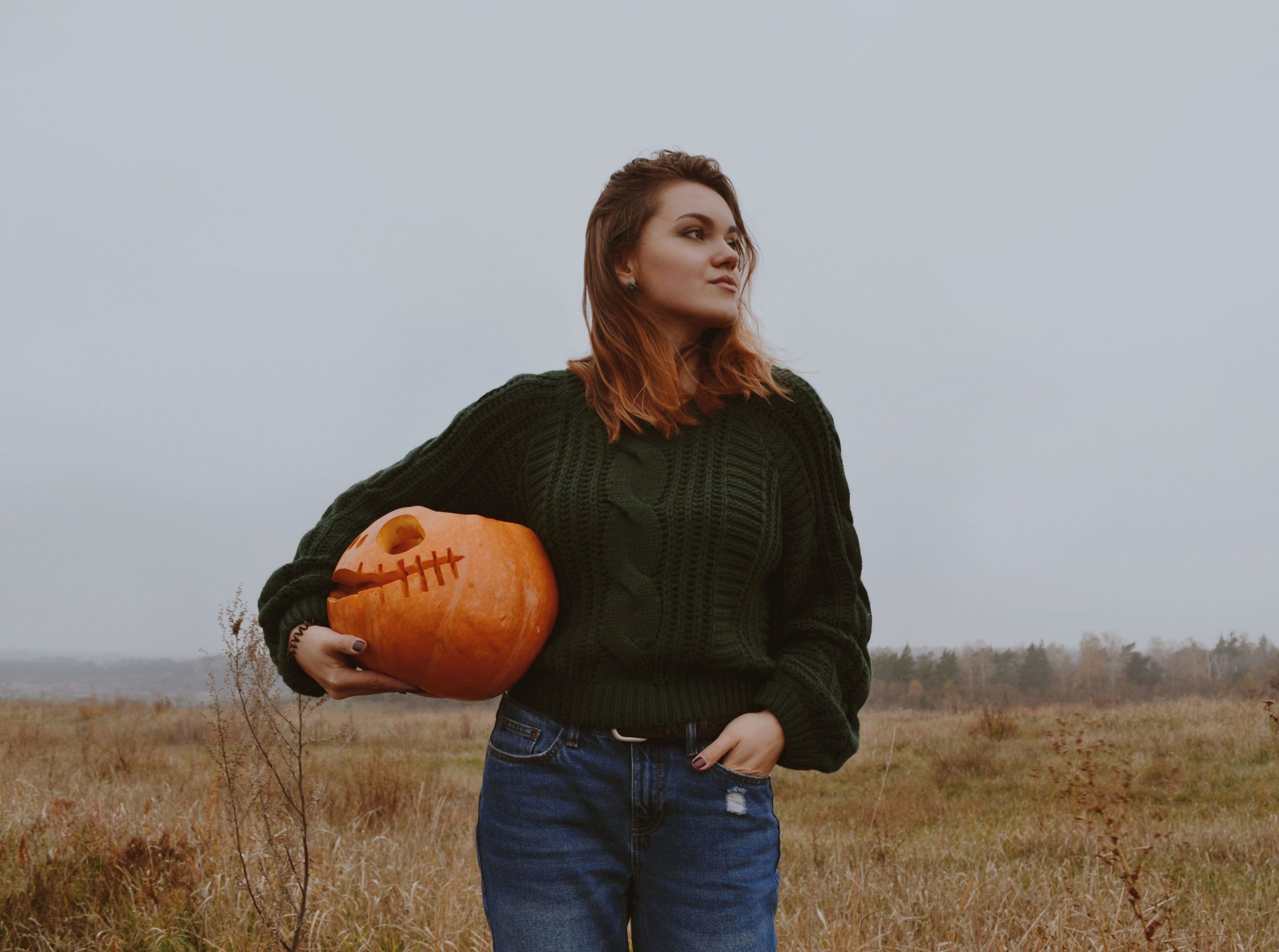 Young woman in green sweater holding jack-o-lantern outdoors in autumn field.