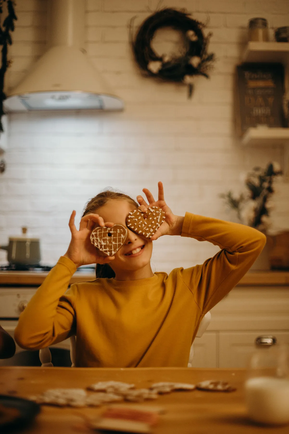 A cheerful girl playfully holds heart-shaped Christmas cookies in a cozy kitchen setting.