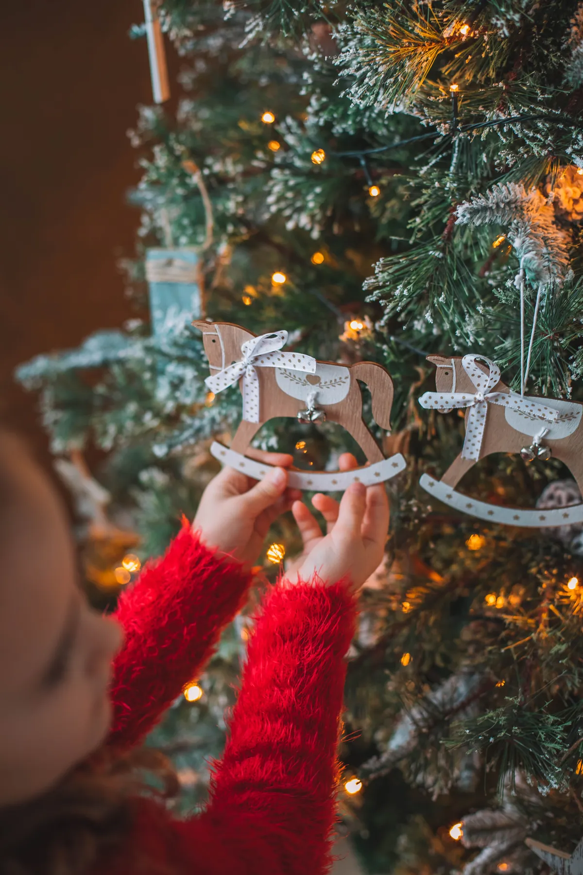 A child in a red sweater decorates a Christmas tree with wooden horse ornaments indoors.