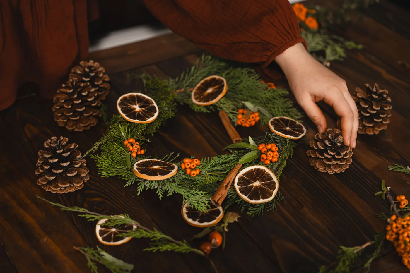 A close-up of a person arranging a Christmas wreath with pinecones and dried oranges.