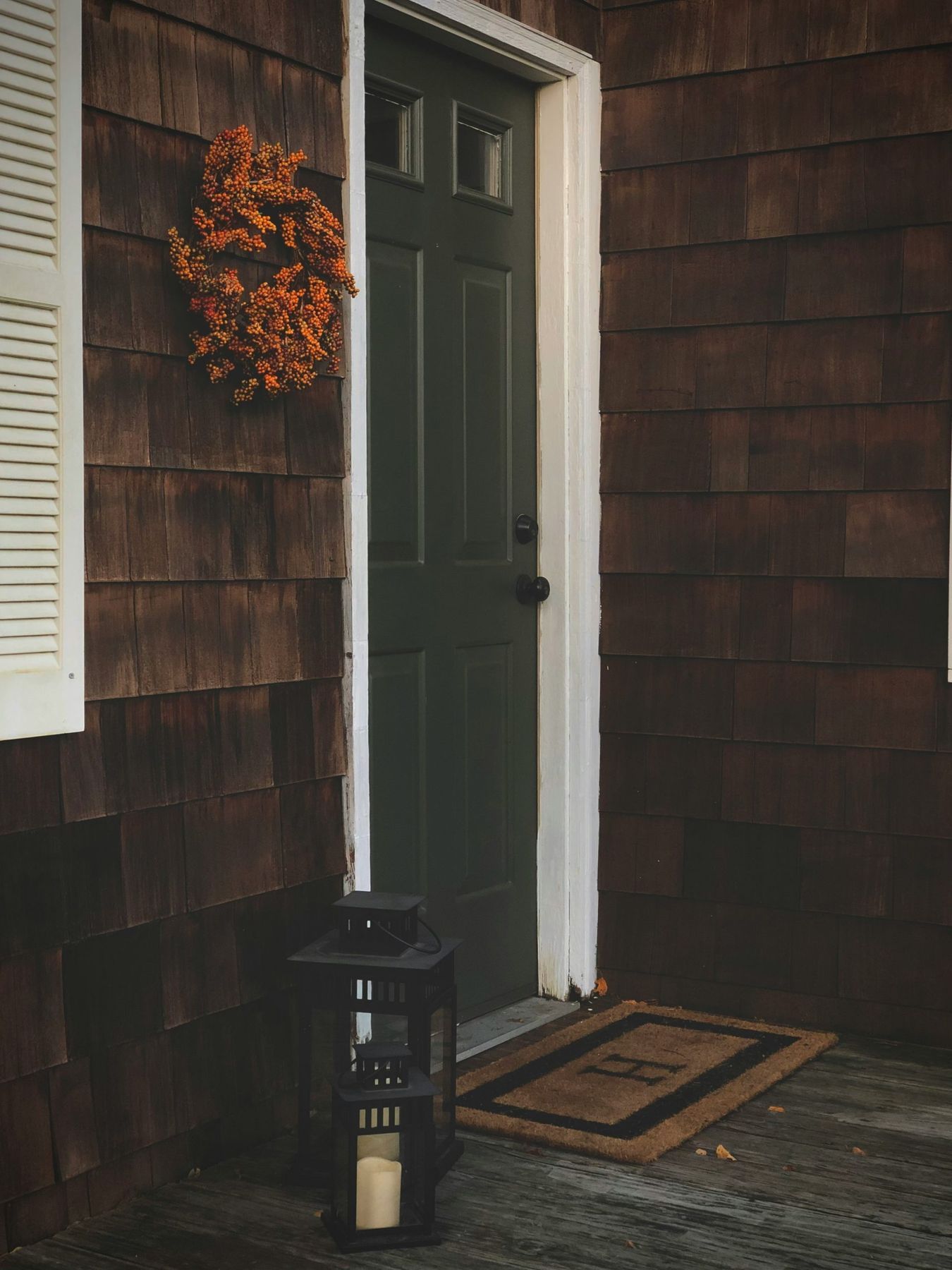 A green door with an orange fall wreath hanging to the left of it. Some black lanterns and a welcome mat with an H sits in front of the door.